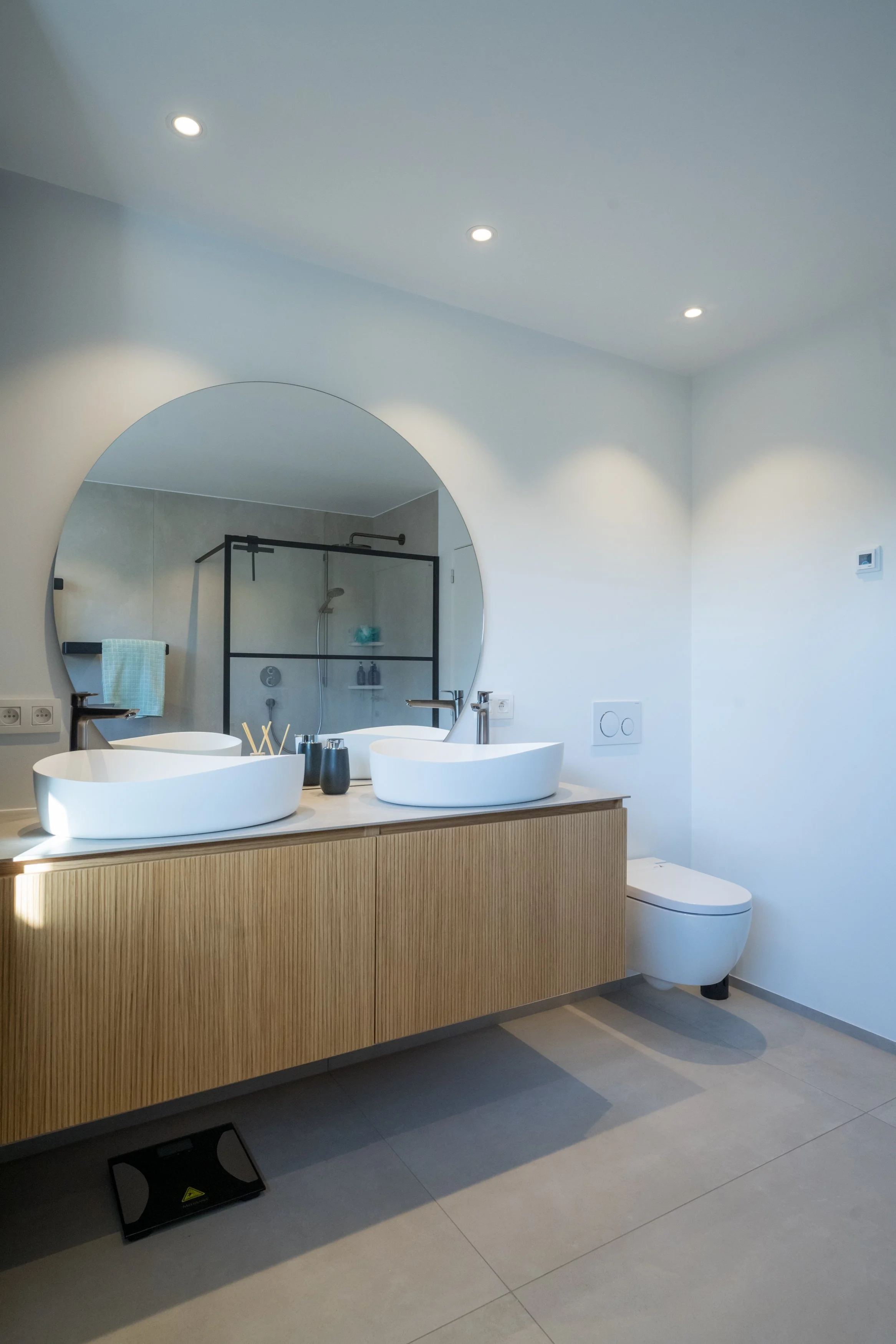 Moderne badkamer met dubbele wit badkuipen op een houten kast onder een grote ronde spiegel, een inloopdouche met glazen wand, witte muren, en een toilet.