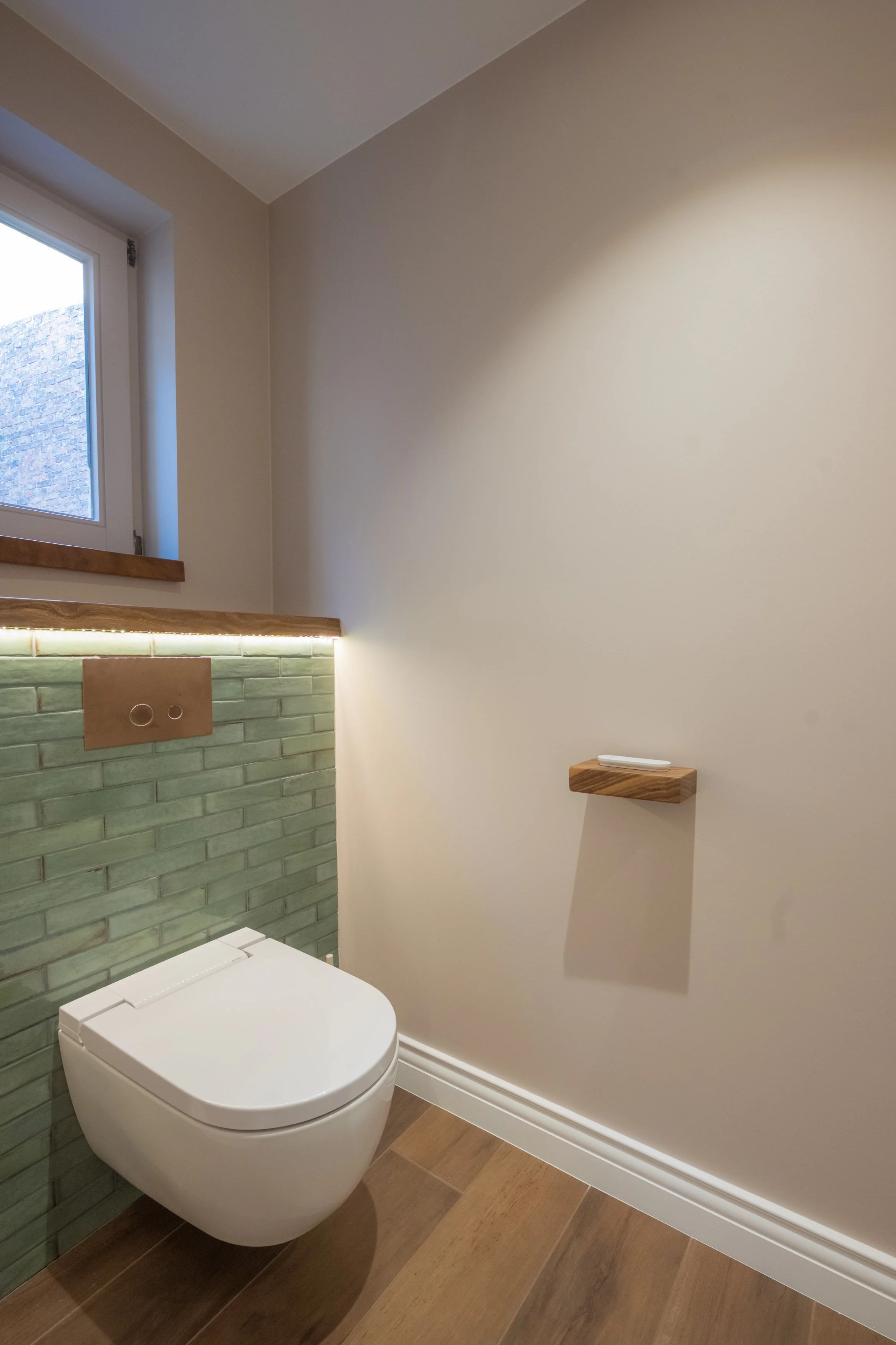 Moderne toiletinrichting met groene bakstenen muur, houten accenten en een klein wandplank.