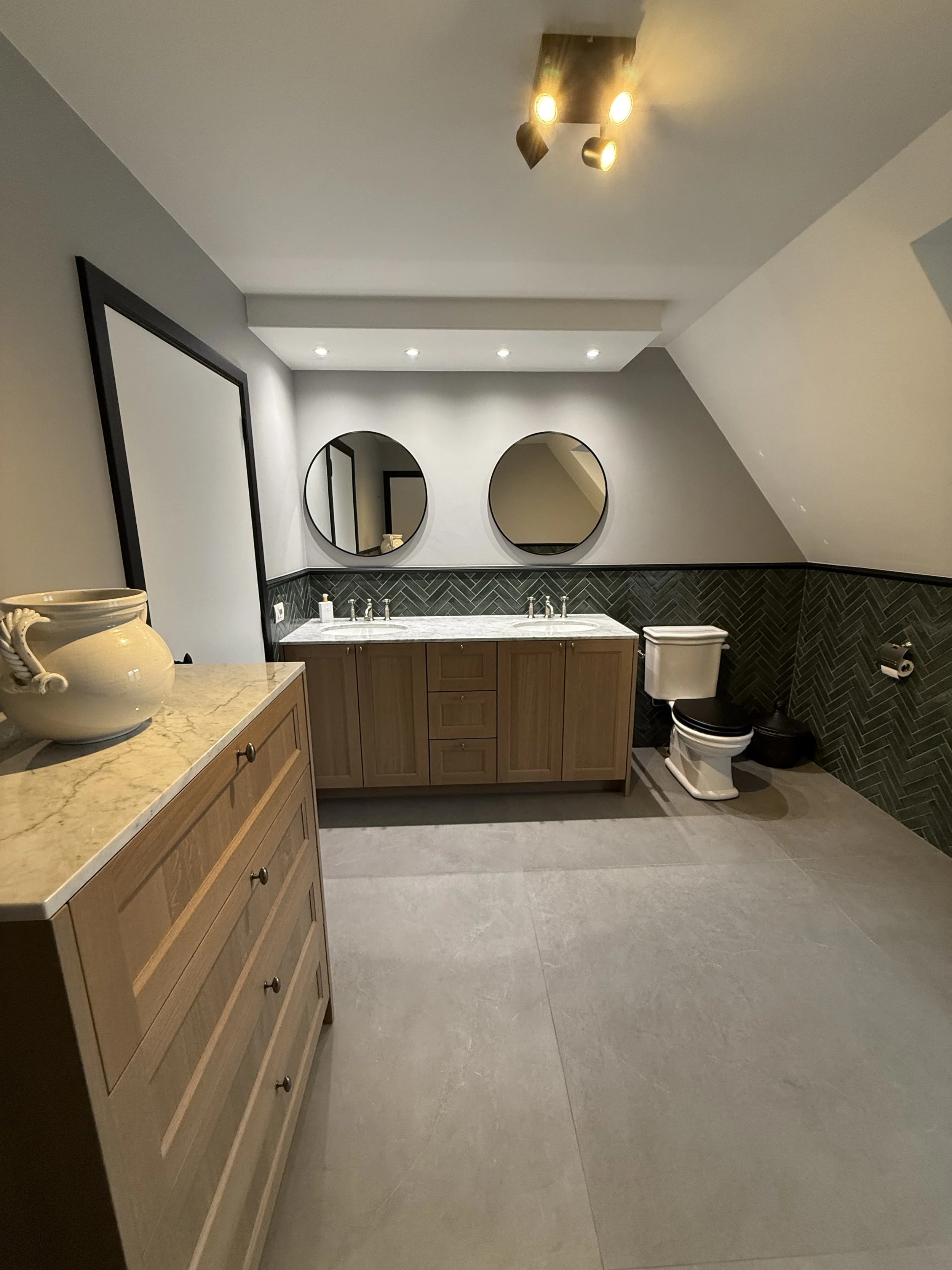 Een modern badkamer met wastafel boven een houten kast, twee ronde spiegels, een toilet, en een plafondlamp met drie richtbare lichten.