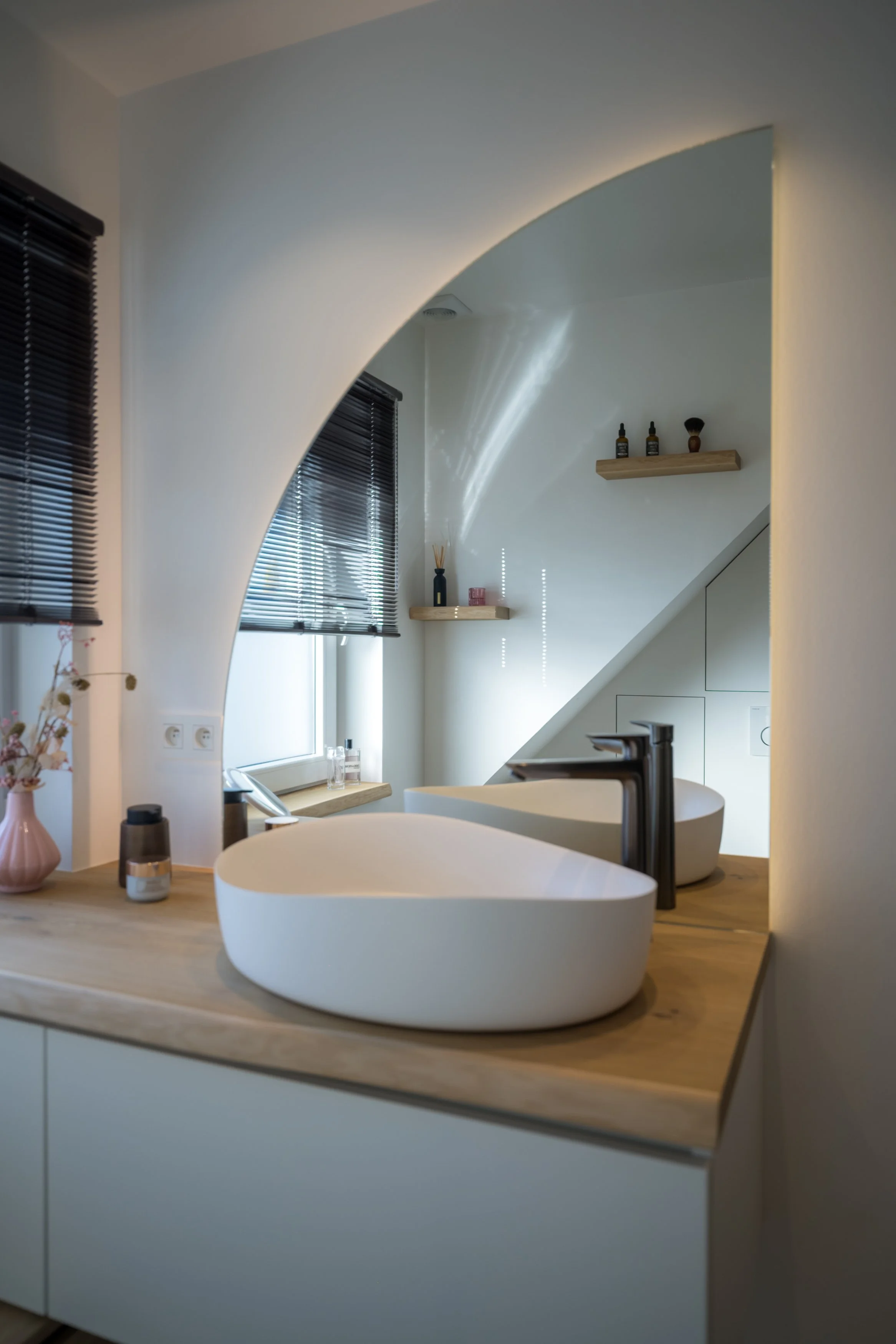 Moderne badkamer met twee witte, ronde wasbakken en zwarte kranen, houten countertop, ramen met zwarte blinds, en een spiegel die de kamer reflecteert.