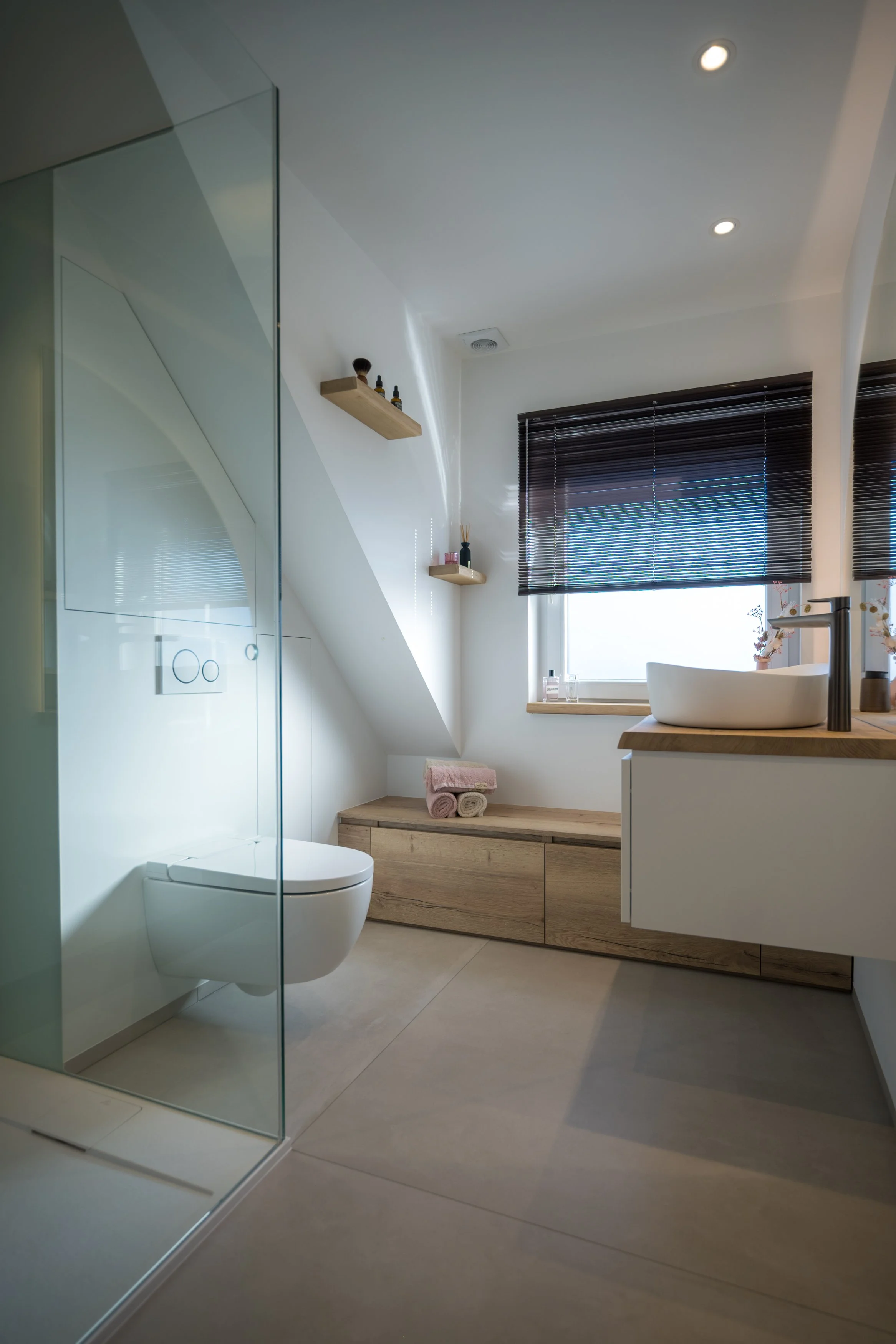 Moderne badkamer met inloopdouche, wandcloset, houten kast, wastafel met wit wasbakje, zwarte raamjaloezieën, en raam met decoratieve bloemen.