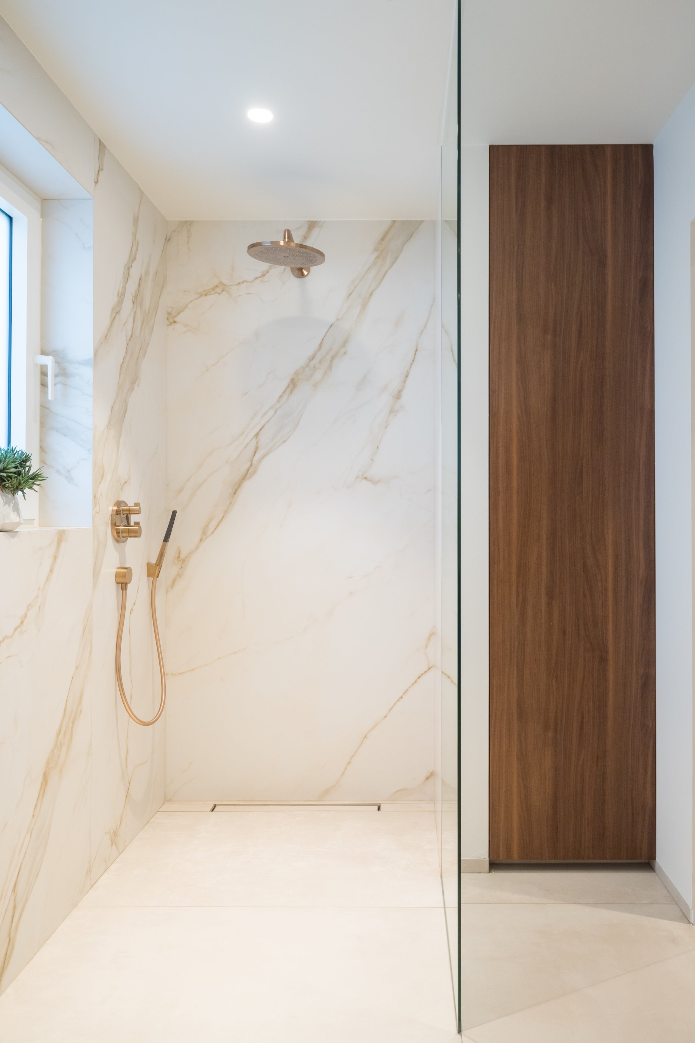 Moderne inloopdouche met witte marmeren wand en gouden douchekranen, houten kast en kleine raam met plant, in een lichte badkamer.