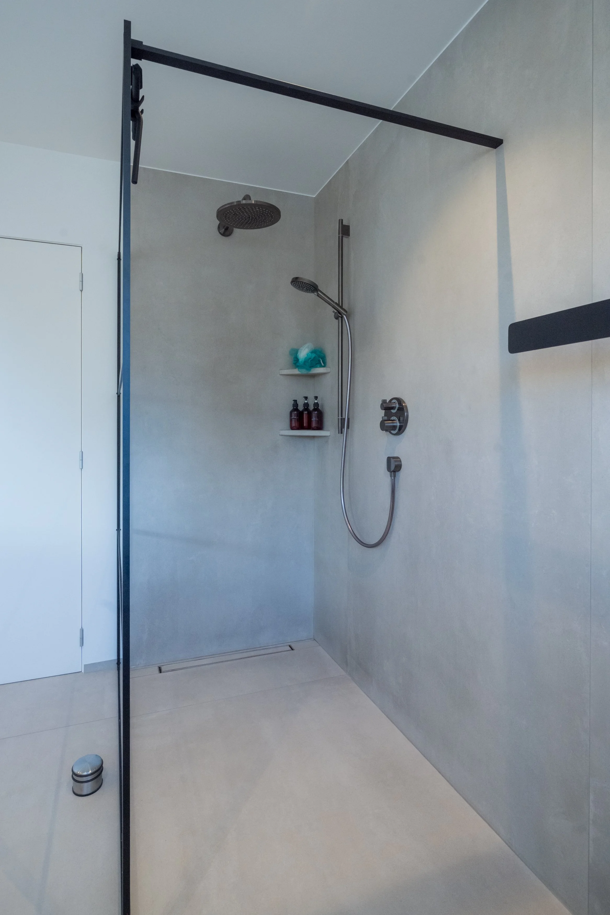 Moderne inloopdouche met twee regen- en handdouches, wandgemonteerde kranen, twee witte plankjes met zeep en shampoo, doorzichtige scheidingswand, beige vloer en muur met een strakke afwerking.