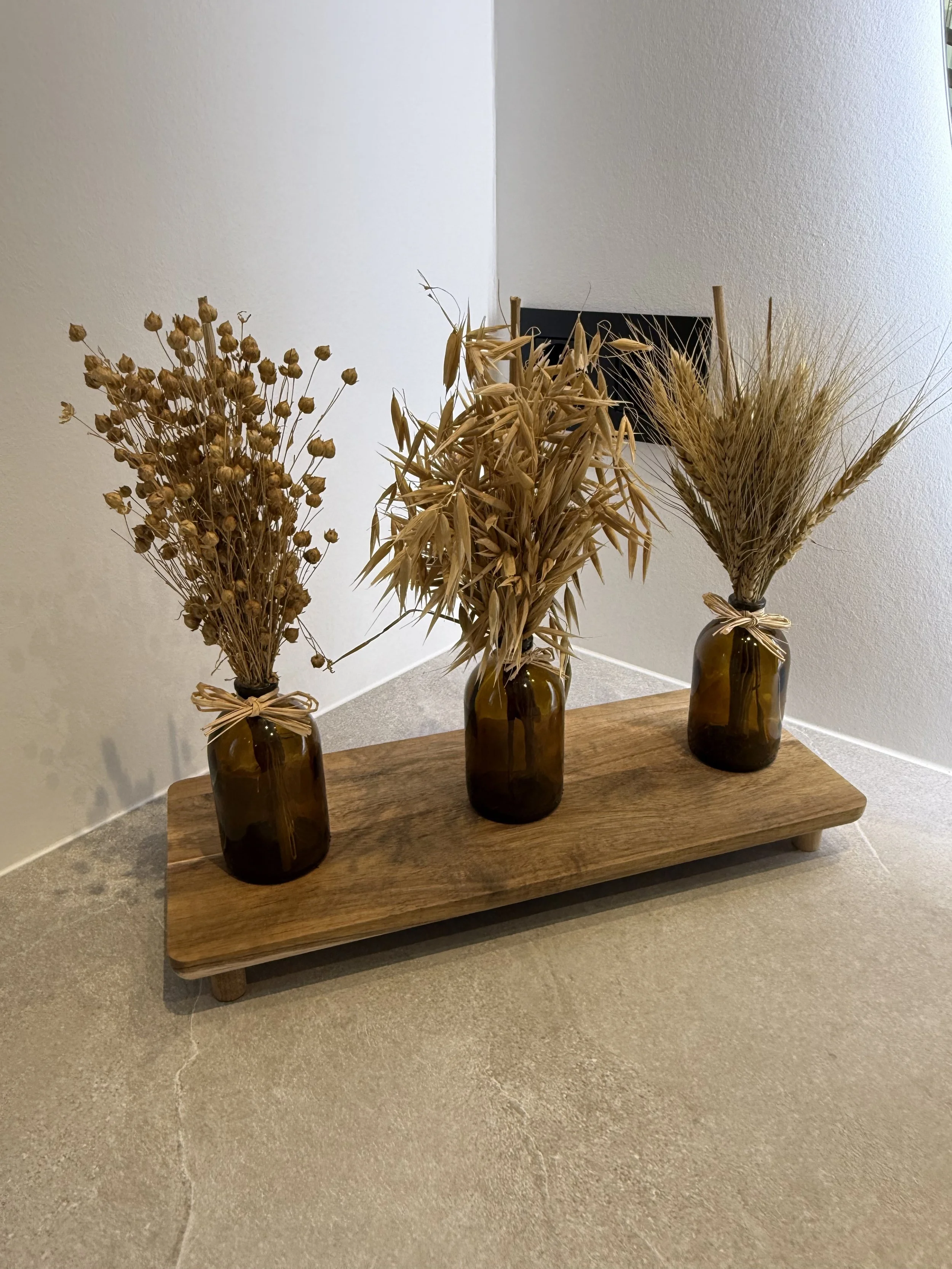 Drie kunstboeketten van droogbloemen in bruine glazen vazen op een houten dienblad op de vloer.