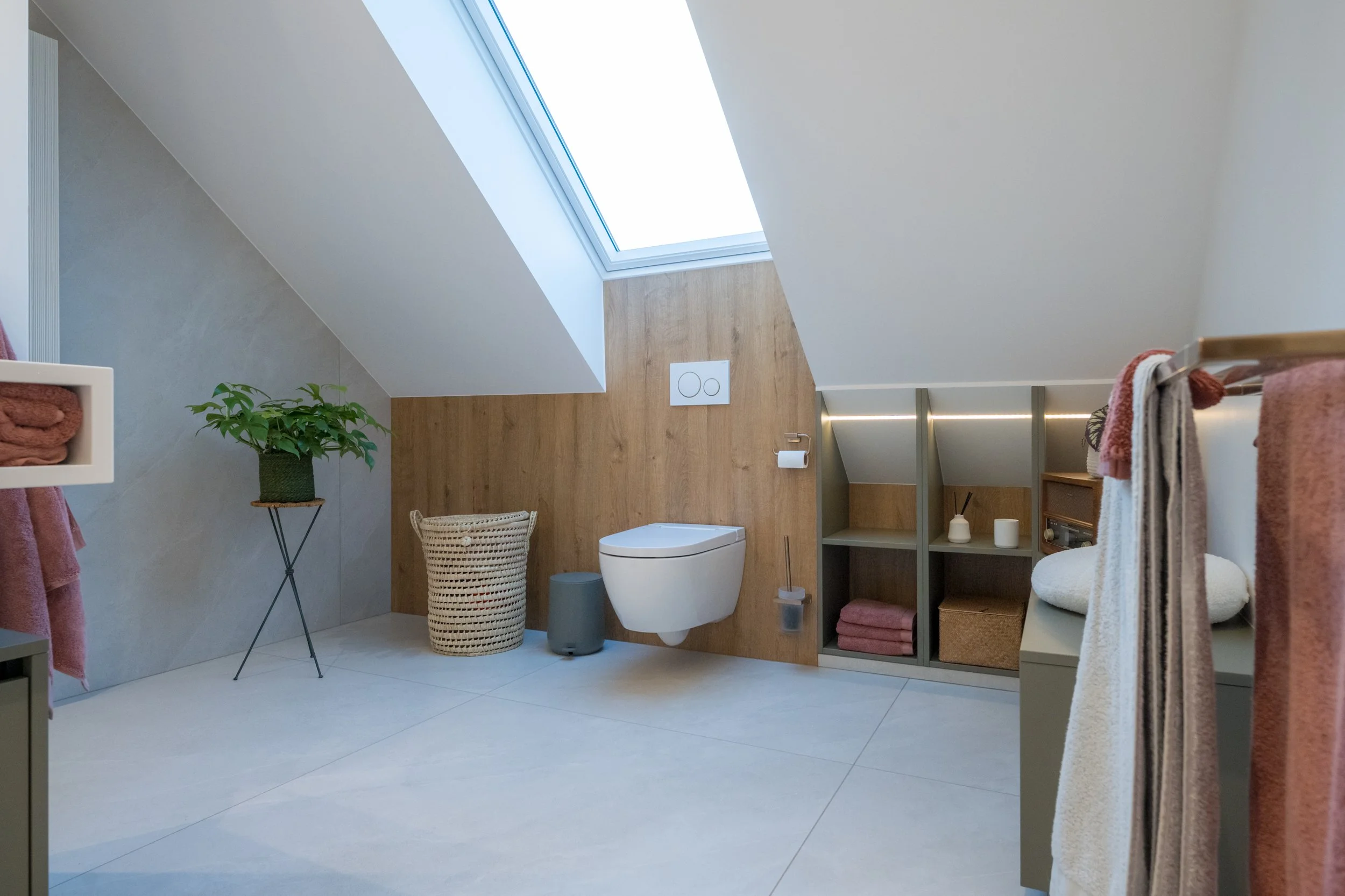 Moderne badkamer met een plafondlichtkoepel, een hangtoilet, houten en grijze wandafwerkingen, een wasrek met handdoeken, een plantenpot, en open planken met handdoeken en decoraties.