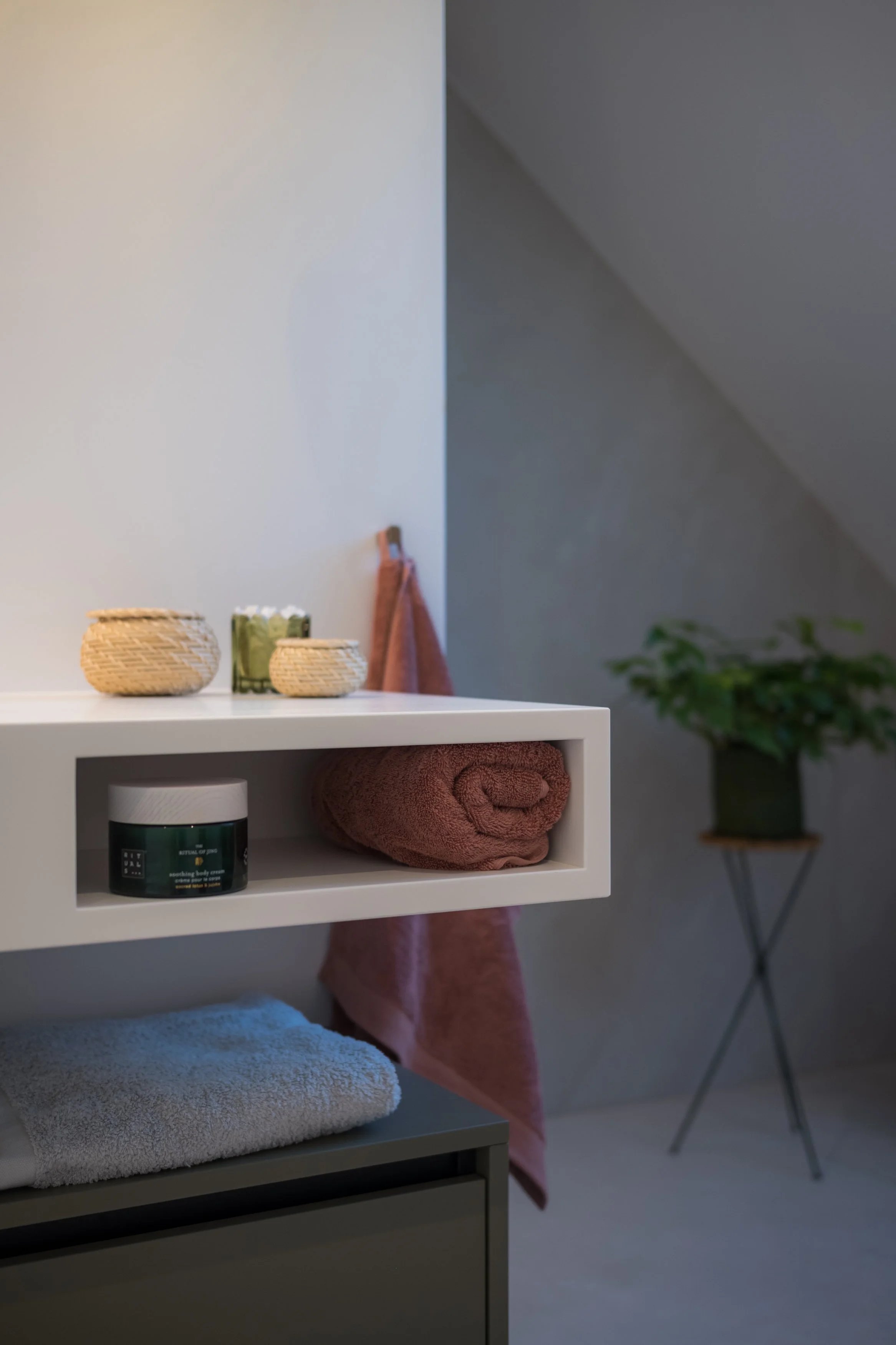 Een wit badkamermeubel met handdoeken, huidverzorgingsproducten en decoratieve items, een groene plant op een metalen standaard, en een muur met een hoek onder een zacht licht.