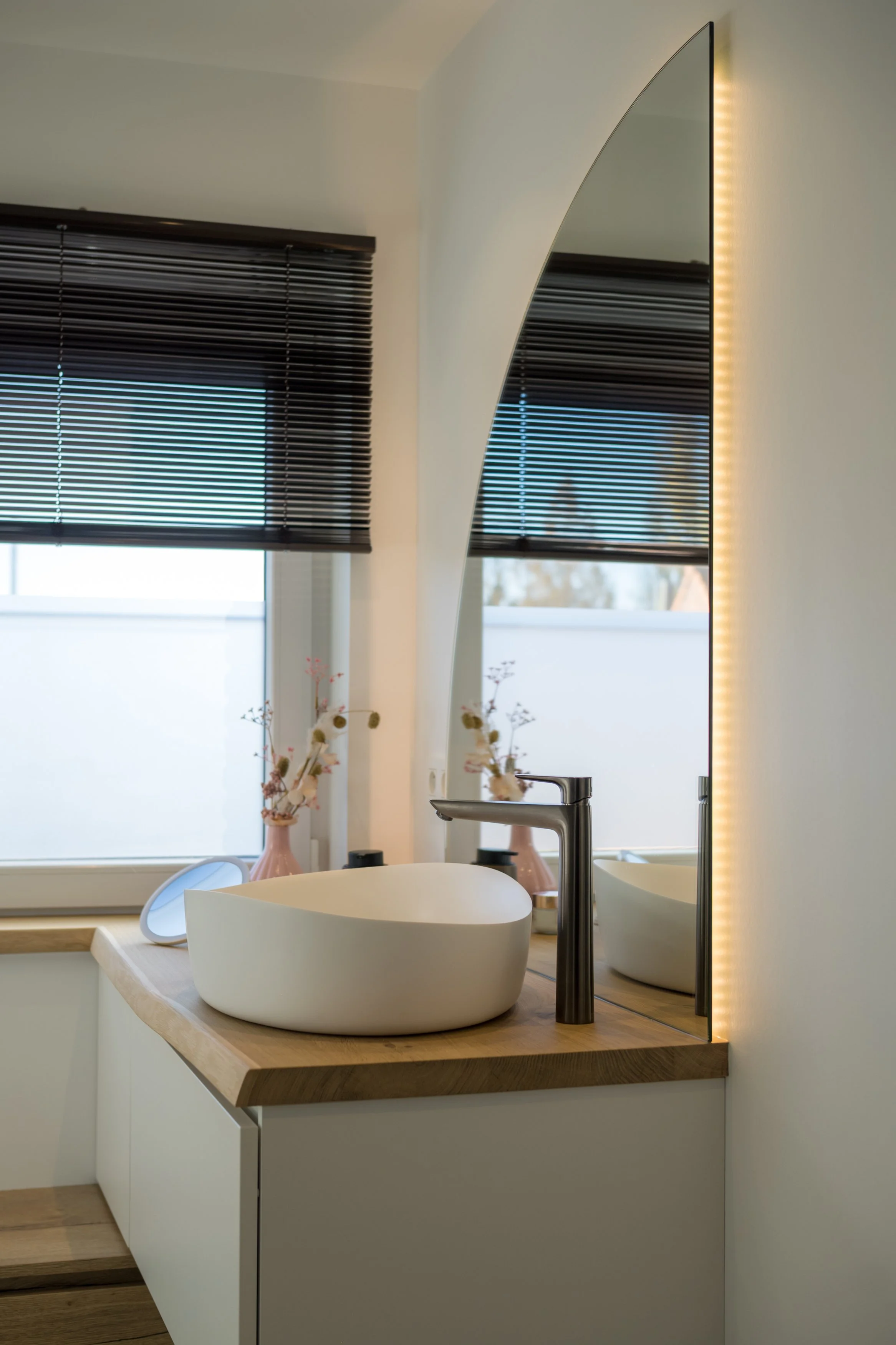 Moderne badkamer met een schelpvormige witte wastafel, een spiegel met LED-verlichting, een raam met zwarte jaloezieën en twee vaasjes met gedroogde bloemen op een houten wastafel.