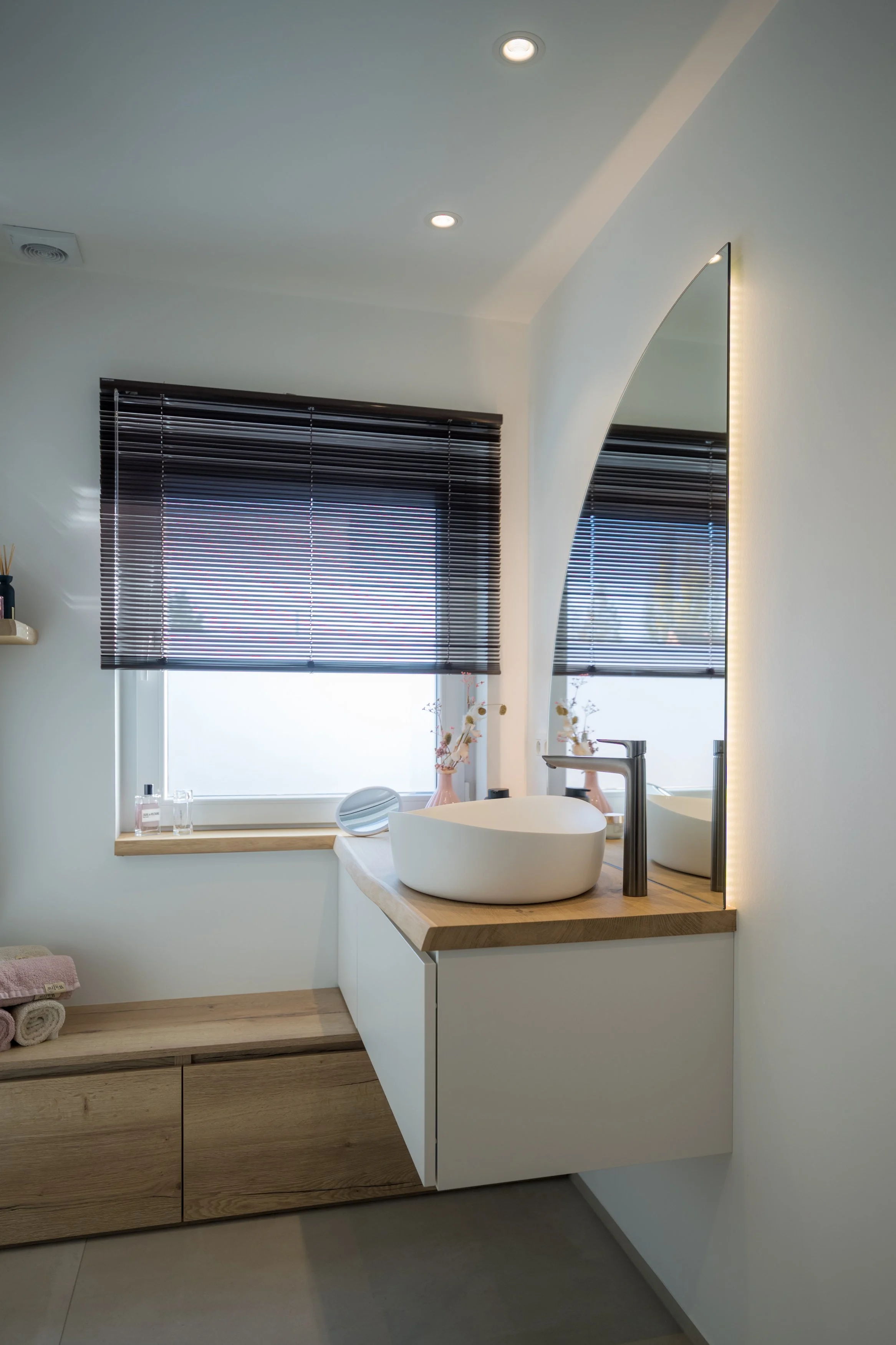 Moderne badkamer met witte wand, houten en witte kast, wastafel met zwarte kraan, spiegel met geïntegreerde verlichting, raam met zwarte jaloezieën, decoratieve vazen en handdoeken.