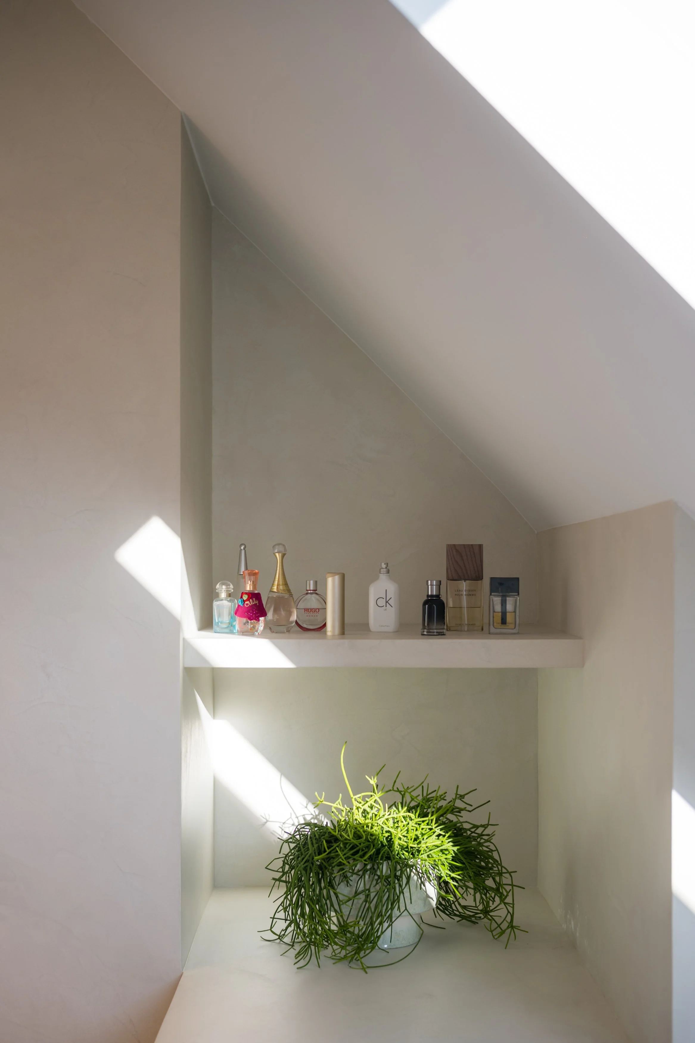 Ingericht nis met diverse parfumflesjes en een groene plant onder een schuin plafond met zonlicht dat binnenvalt.