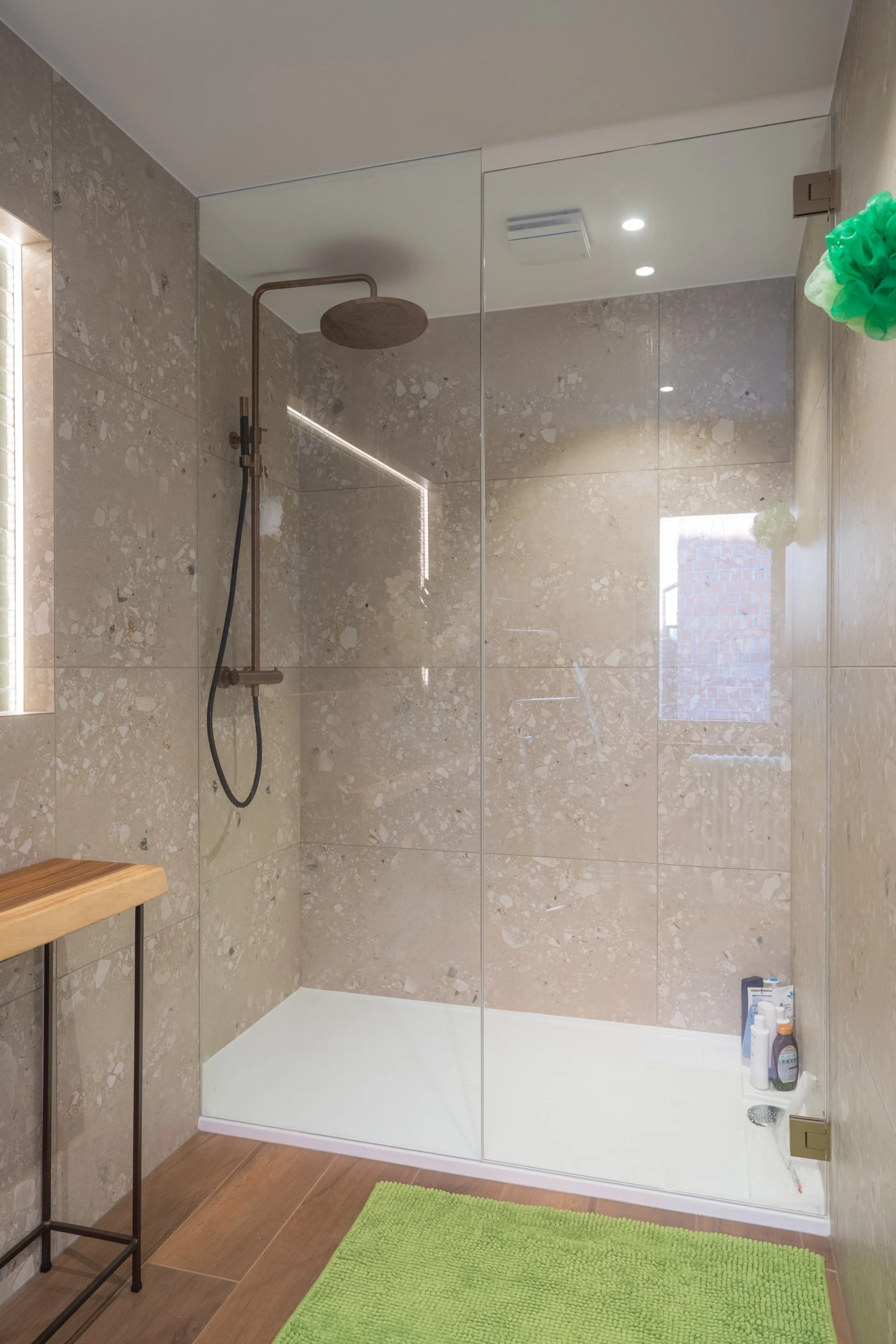 Binnen in een moderne inloopdouche met beige natuurstenen tegels, een houten plankje, een groen badmat en diverse badproducten in de hoek.
