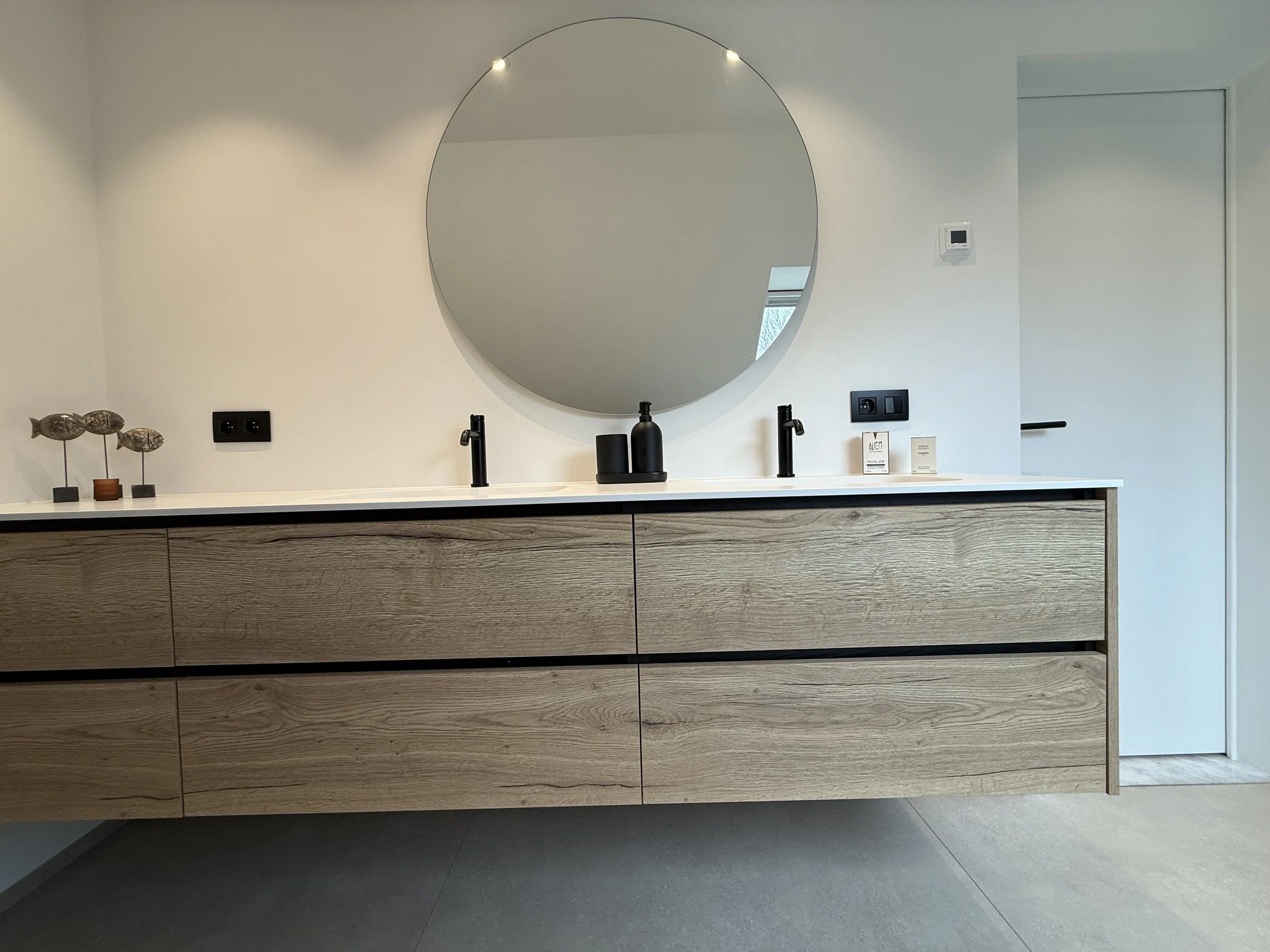 Moderne badkamer met houten kast, dubbele wastafels, ronde spiegel, zwart kranen, en decoratieve elementen.