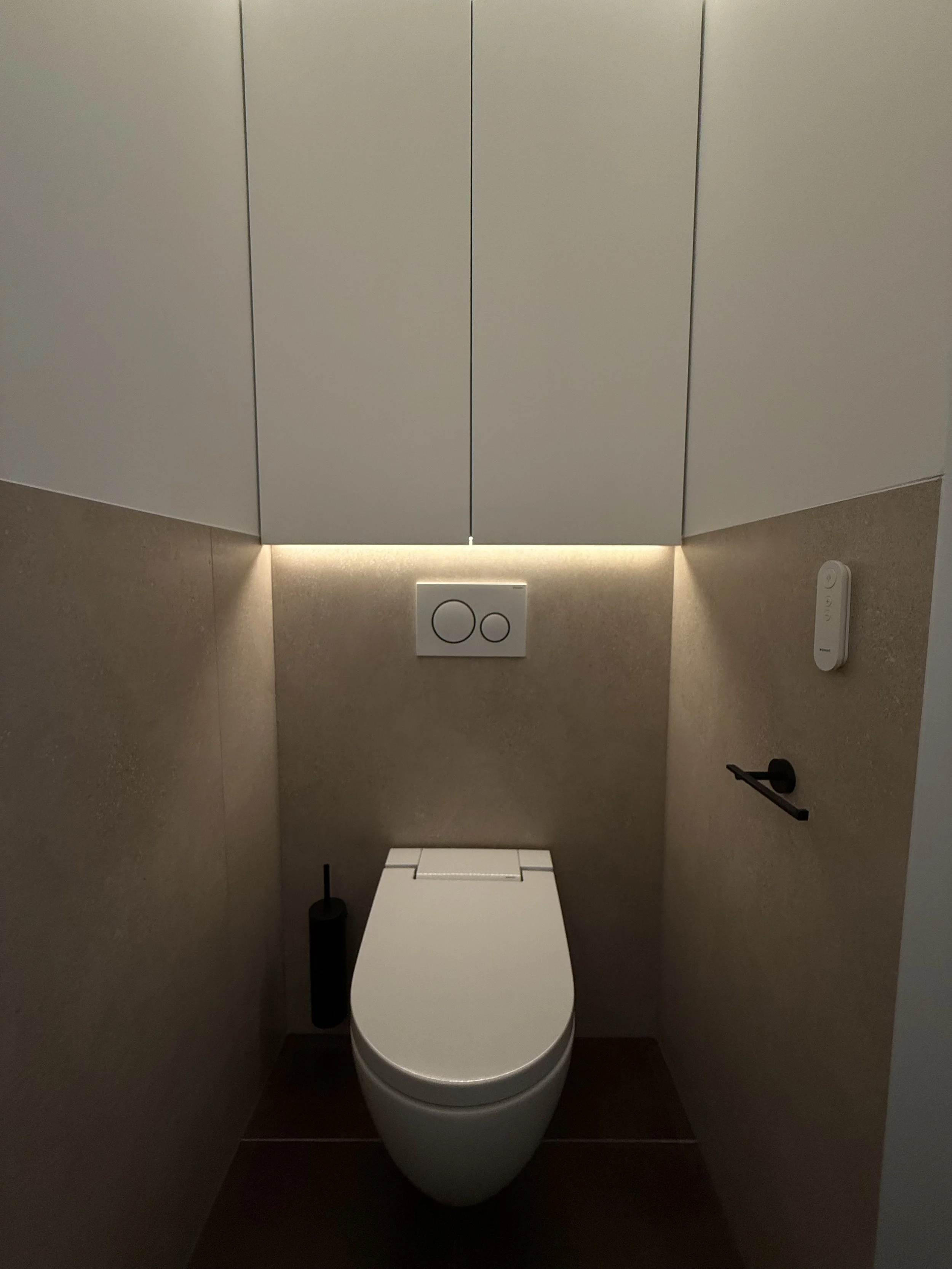 Een modern toilet met een wit toiletbril, een toiletrolhouder, een toiletborstel, een inbouwtoilet met een wandmodel bedieningspaneel, en bovenkasten met verlichting in een kleine badkamer.