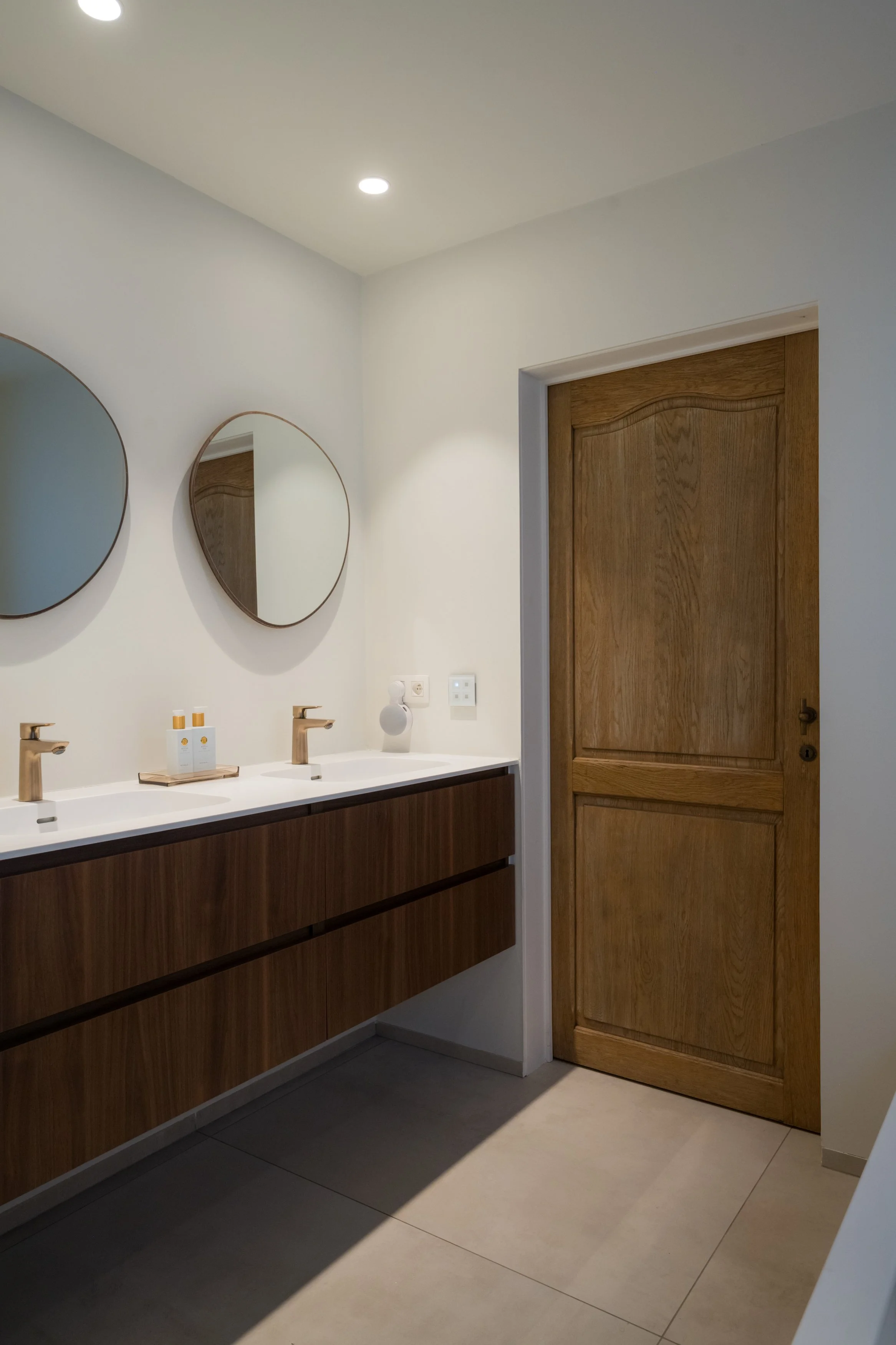Badkamer met dubbele wastafel, twee ronde spiegels, houten kast onder de wastafel, houten deur, witte muur, plafondlamp, contactdoos en zeepbakje.