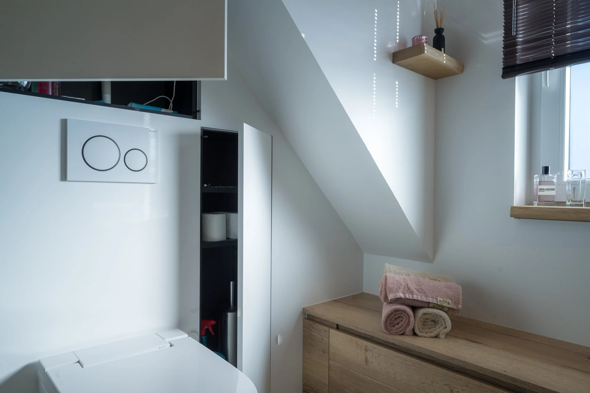 Een wasruimte met een wasmachine, een witte kast, open muurplanken met decoratieve voorwerpen, een venster met donkerbruine jaloezieën, en gestapelde handdoeken op een houten meubel.