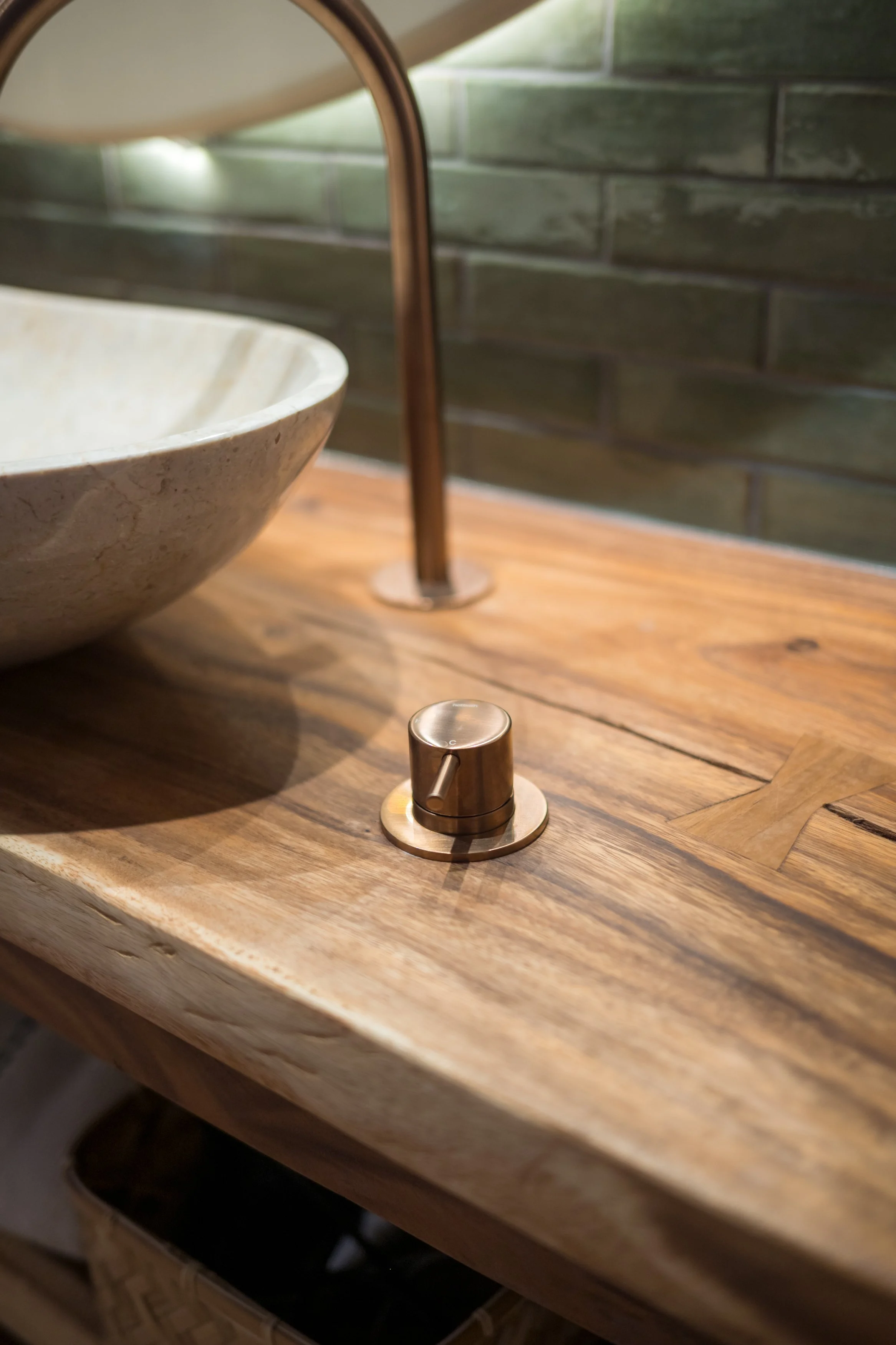 Koperkleurige kraan op houten wastafel met een waskom en groene tegels op de achtergrond.