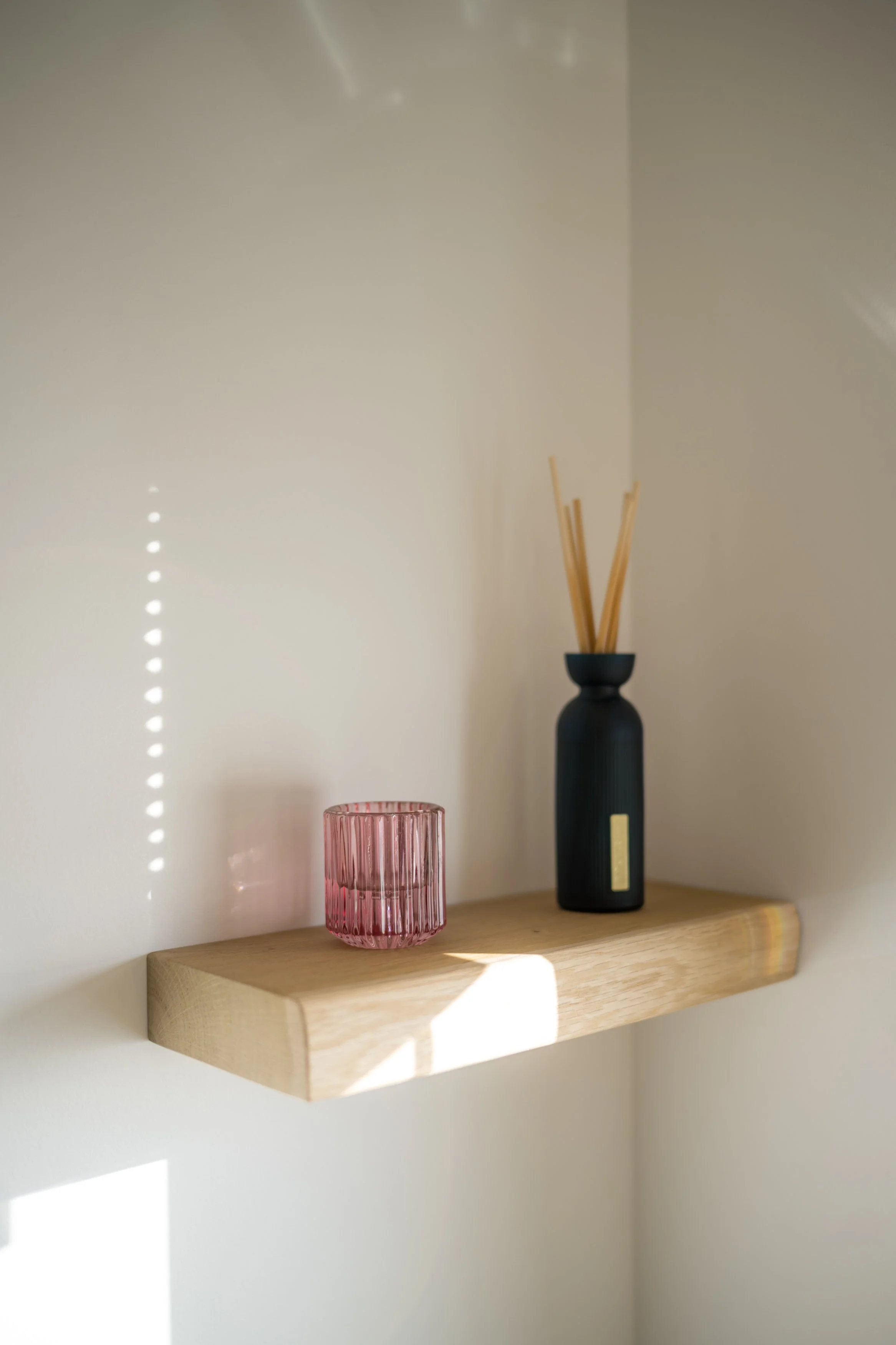 Een houten plank met een roze glazen en een zwarte vaas met riet. Ner- en schaduw op een witte muur.