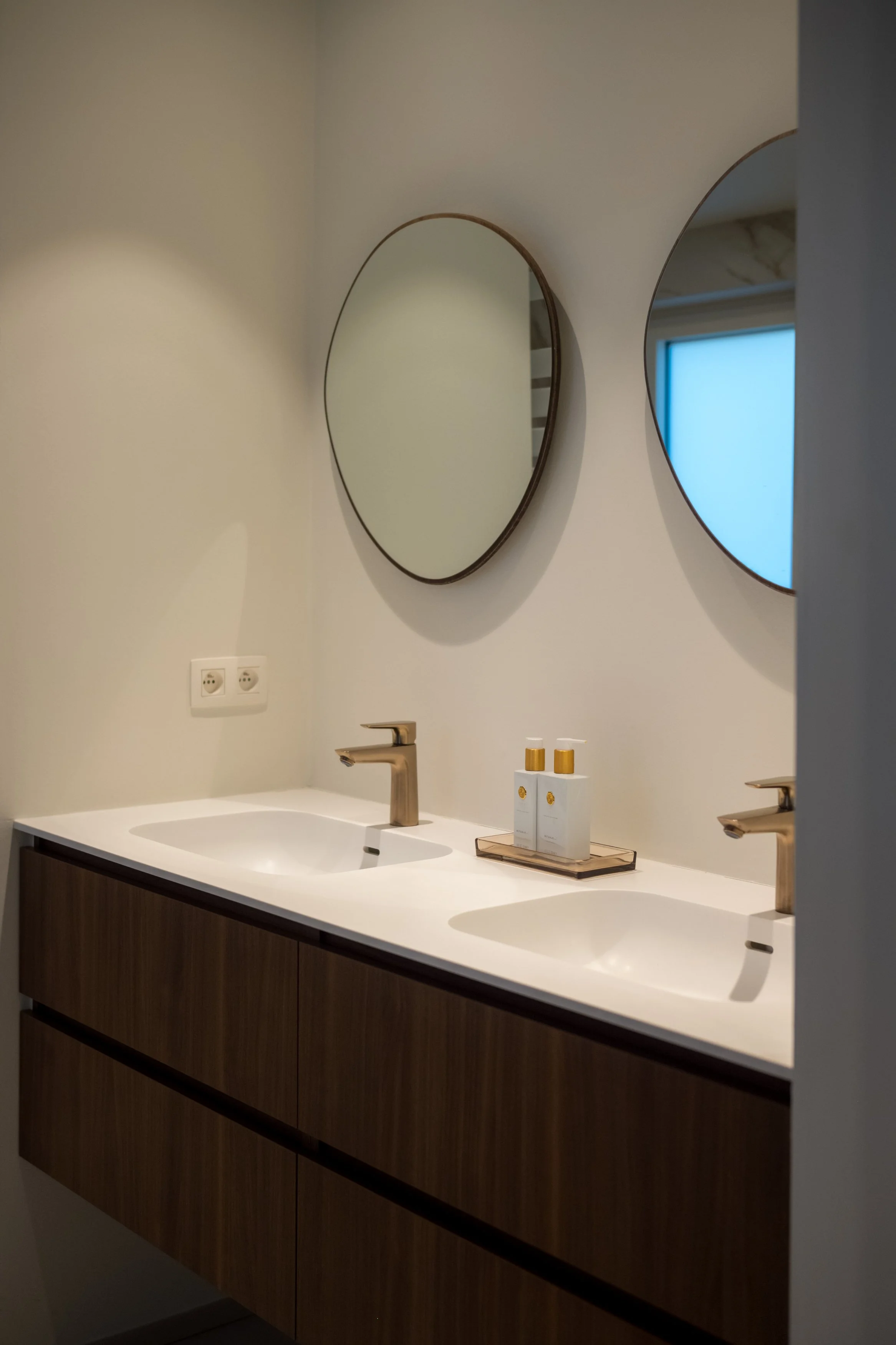 Zweibakt badkamermeubel met twee wastafels, gouden kranen, twee ronde spiegels aan de muur, en een natuurlijke houten kast onder de wastafels, met een tray met verzorgingsproducten.