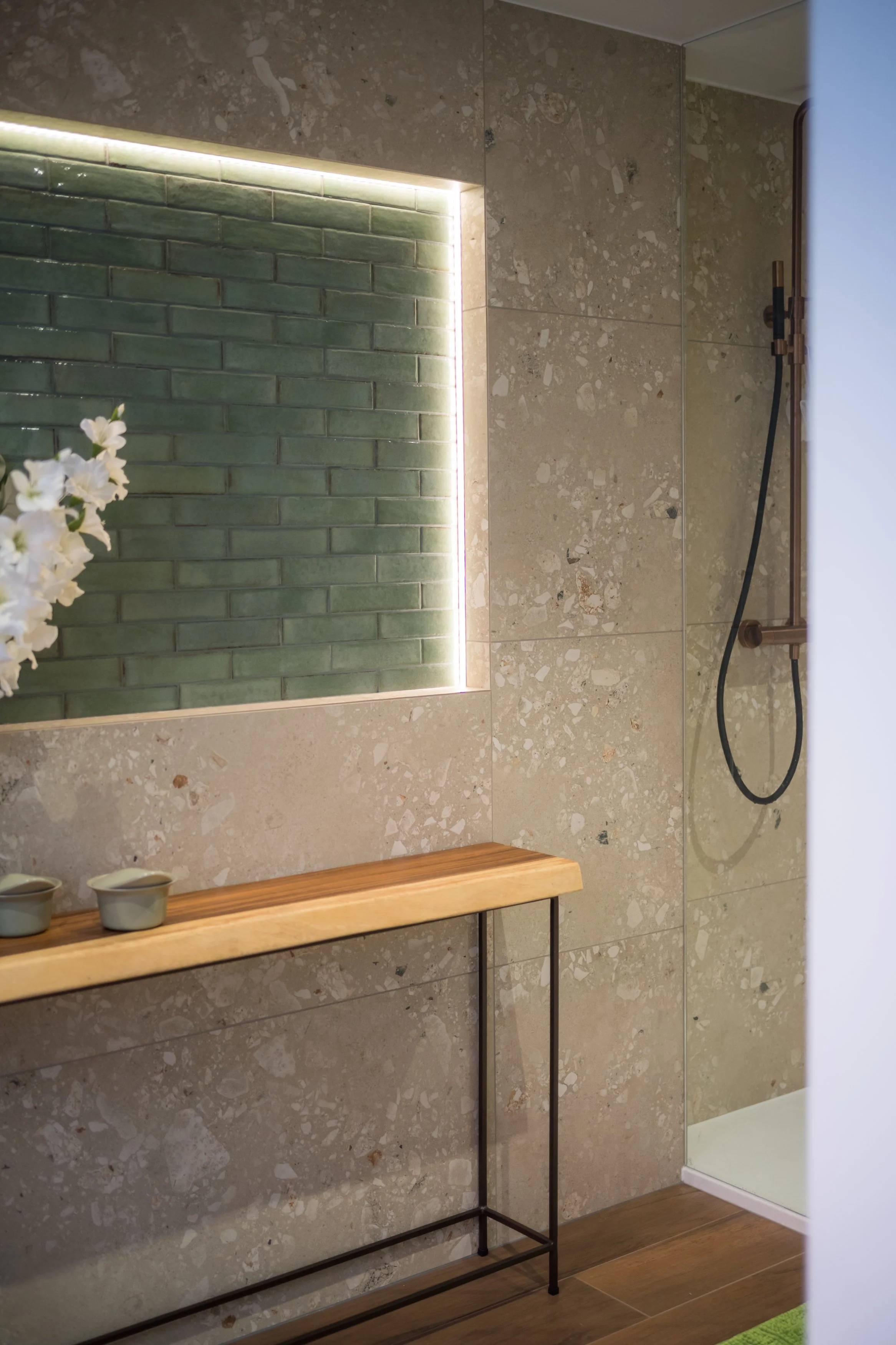 Een moderne badkamer met een inloopdouche, groene bakstenen muur met led-verlichting, een houten plank met twee kleine keramieken kopjes, en beige terrazzo tegels.
