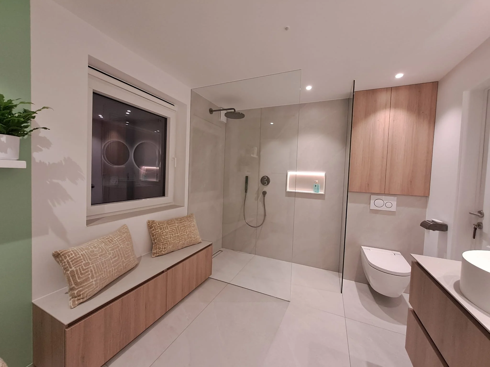 Moderne badkamer met een inloopdouche, een raam met drie waaiervormige patronen in de spiegel en een zitbank met kussens.