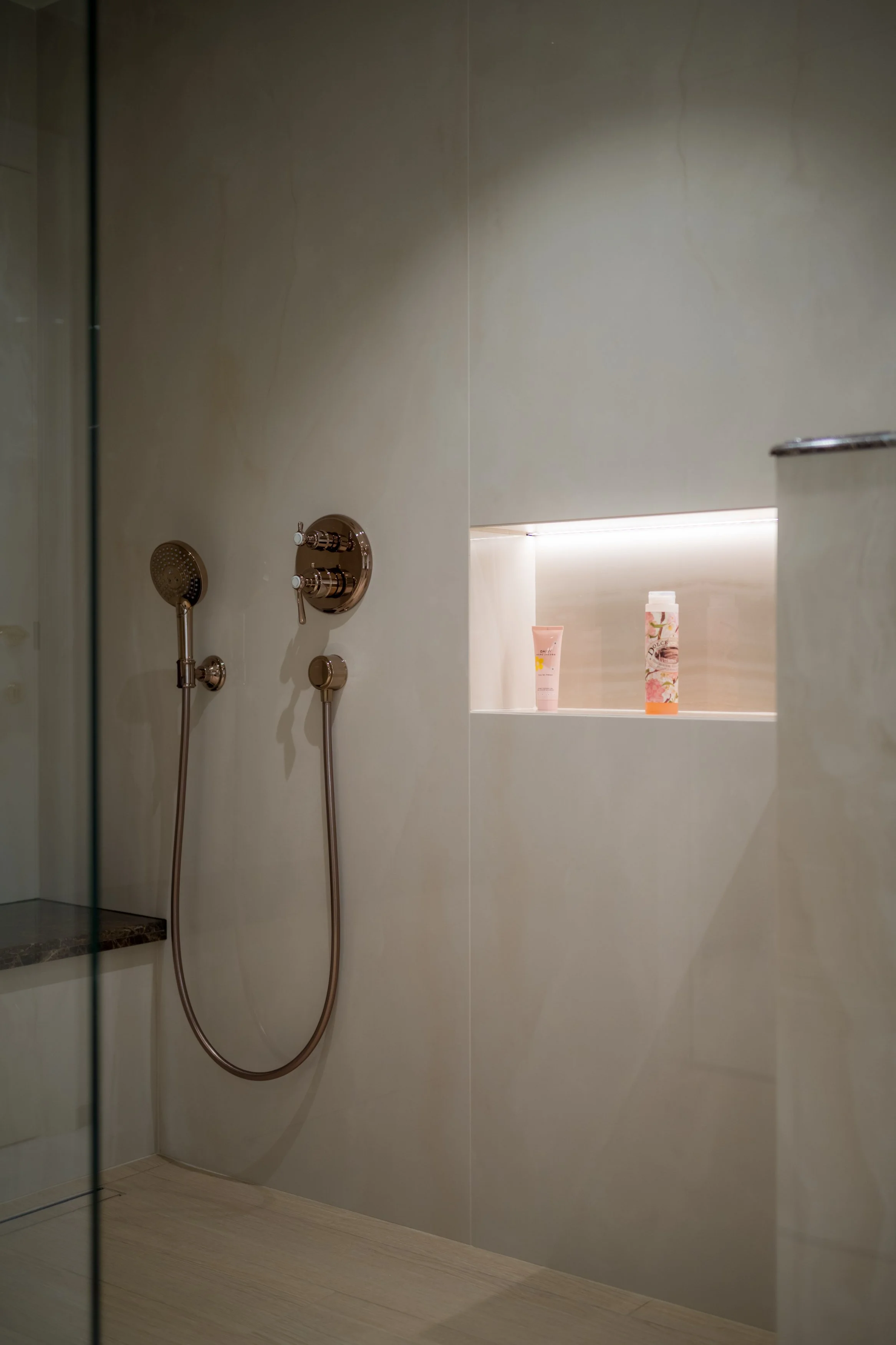 Een moderne inloopdouche met een koperen handdouche en een open ingebouwde nis met verzorgingsproducten.