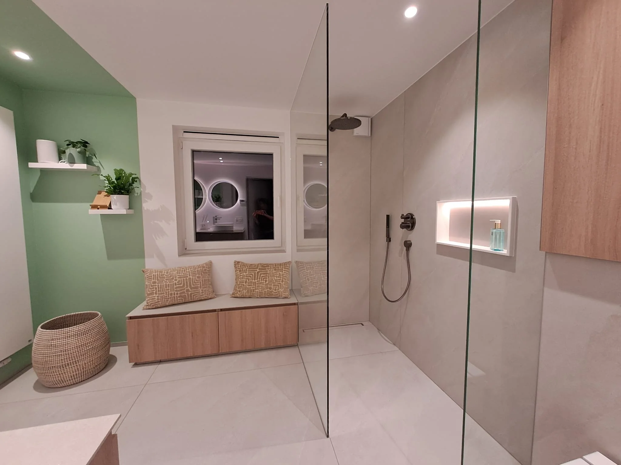 Moderne badkamer met een inloopdouche, een bankje met kussens onder een raam, groene muur met witte planken en planten, en een rieten mand op de vloer.