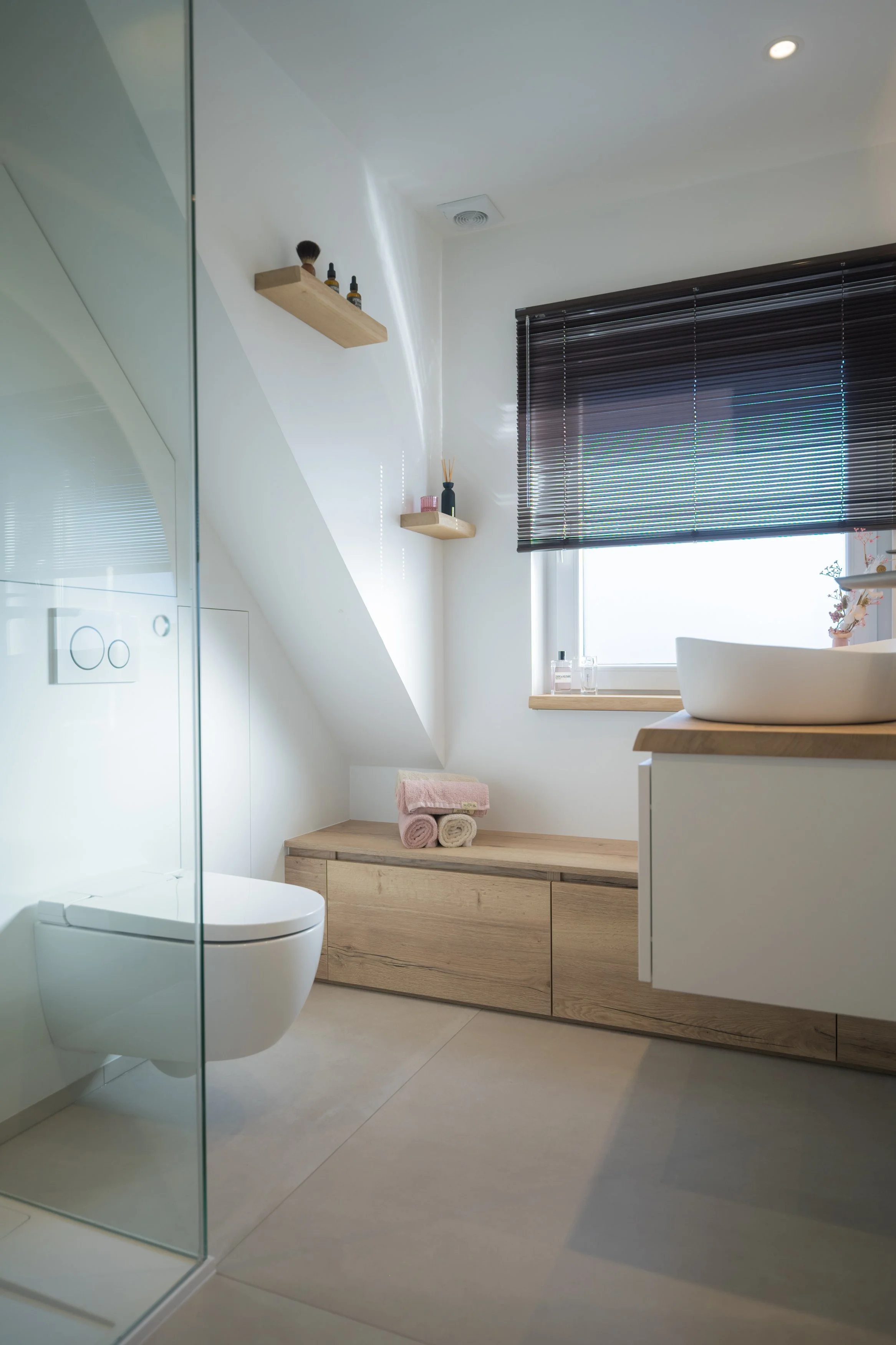 Modern badkamer met toilet, wastafel en donkere raamdecoratie, houten opberglade met handdoeken, ingebouwde bovenkast, en twee wandrekken met decoratieve items.
