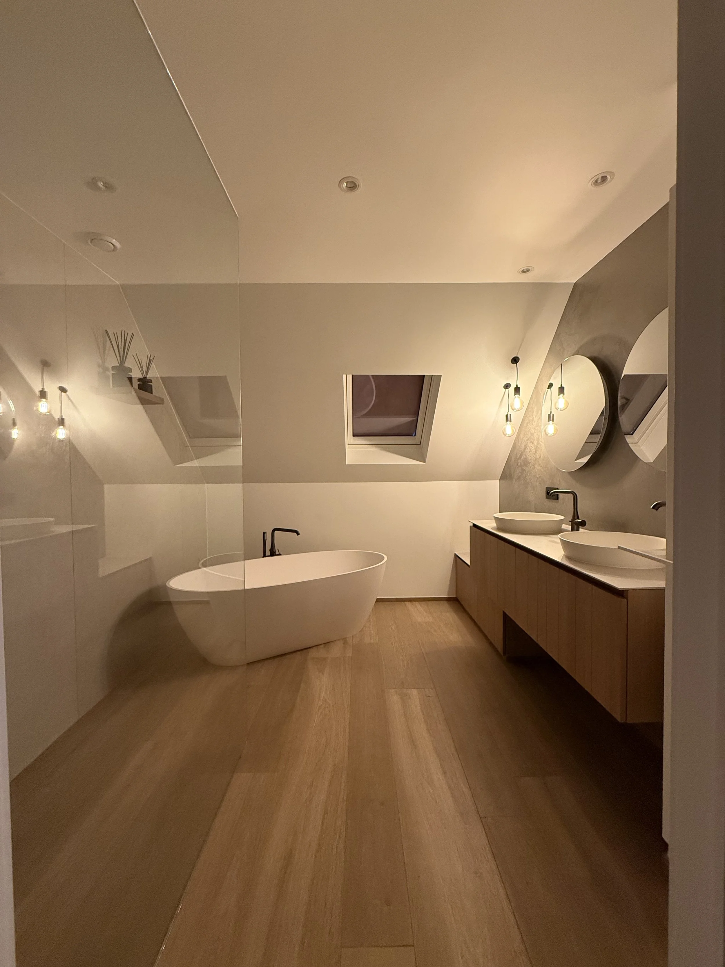 Moderne badkamer met vrijstaand bad, houten vloer, twee witte waskommen met ronde spiegels en hanglampen, dakraam op schuine muur, zacht verlichte sfeer.