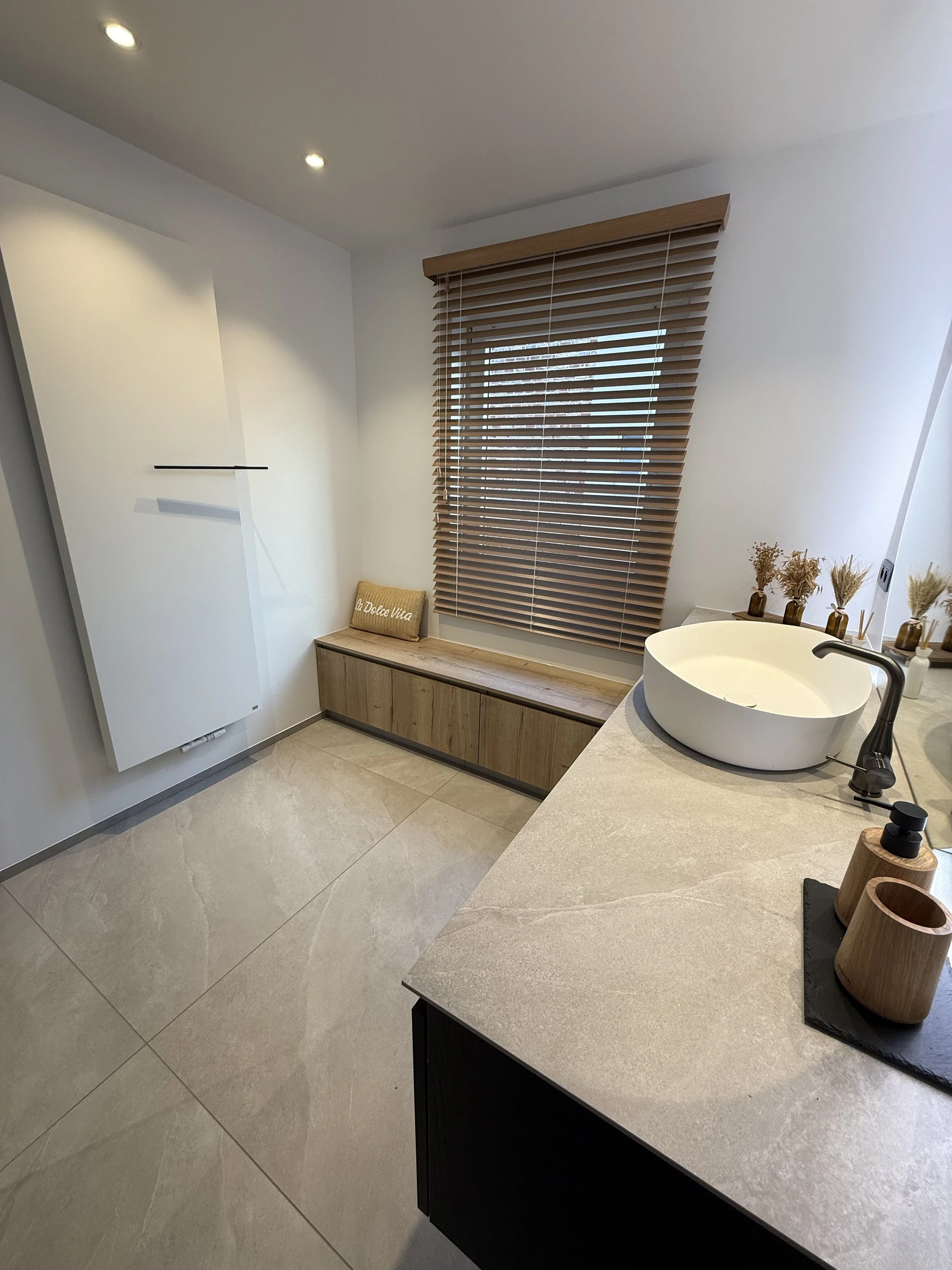 Een moderne badkamer met witte muren, een grote voegloze beige tegelvloer, een raam met houten jaloezieën, een bankje onder het raam en een waskom op het badmeubel naast de spoelbak. Er zijn decoratieve vaasjes met gedroogde bloemen.