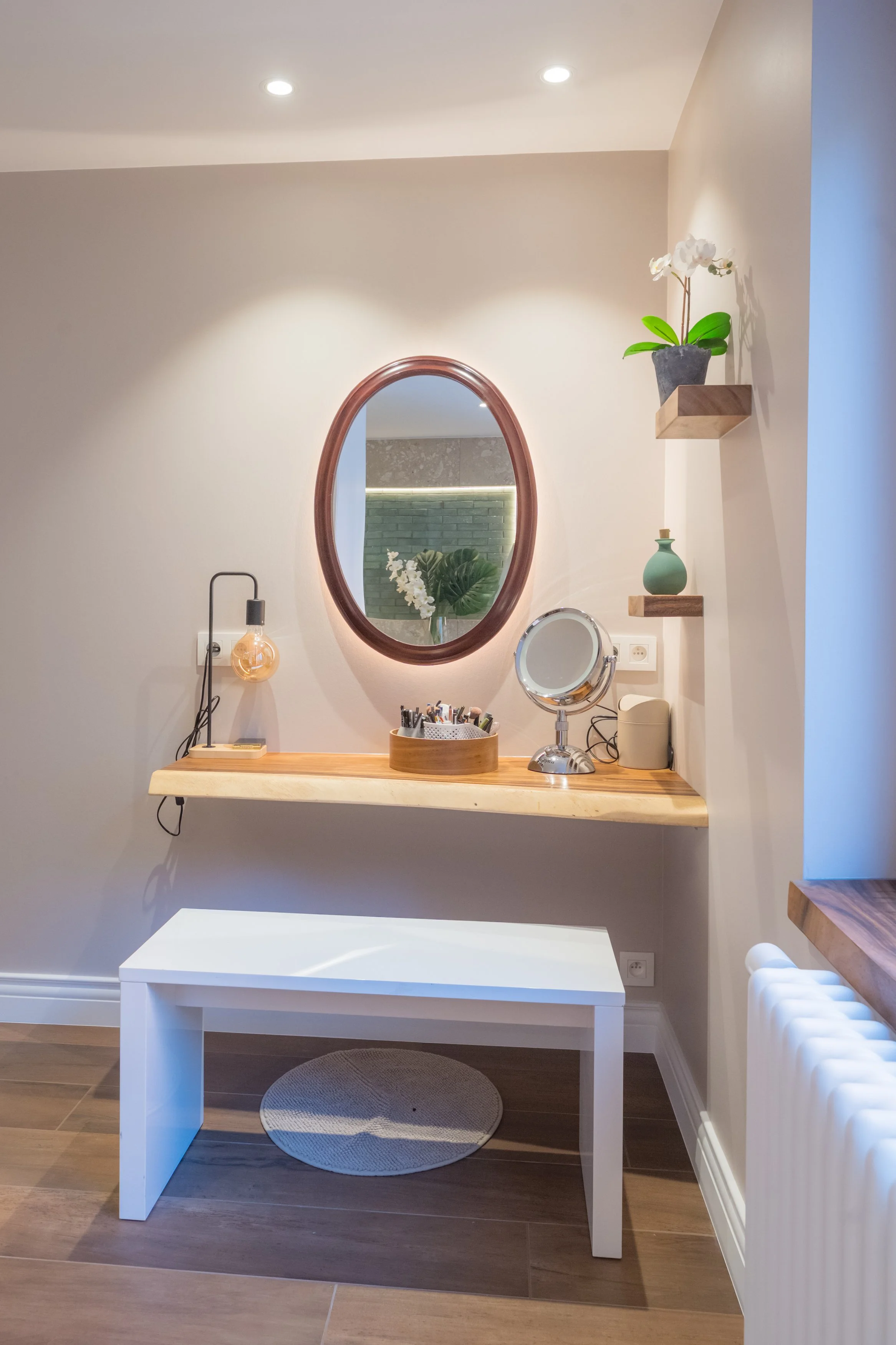 Een spiegel, een bloemstuk, een ronde tafelspiegel, en decoratieve accessoires op een houten tafel, met een witte bank onder de tafel en een radiator aan de rechterkant, alles in een lichte, minimalistische kamer.