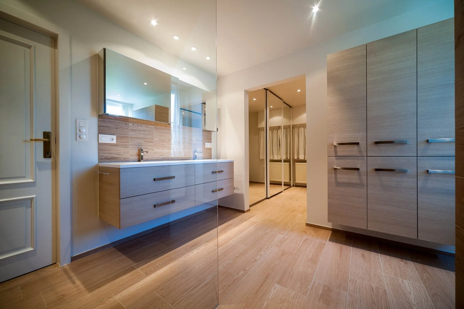 Modern badkamer met grote spiegel, houten kasten en vloer, wastafel met dubbele kraan en een spiegelwand, lichte muren en een deur, tevens een extra ruimte met grote kast en een raam met gordijnen.