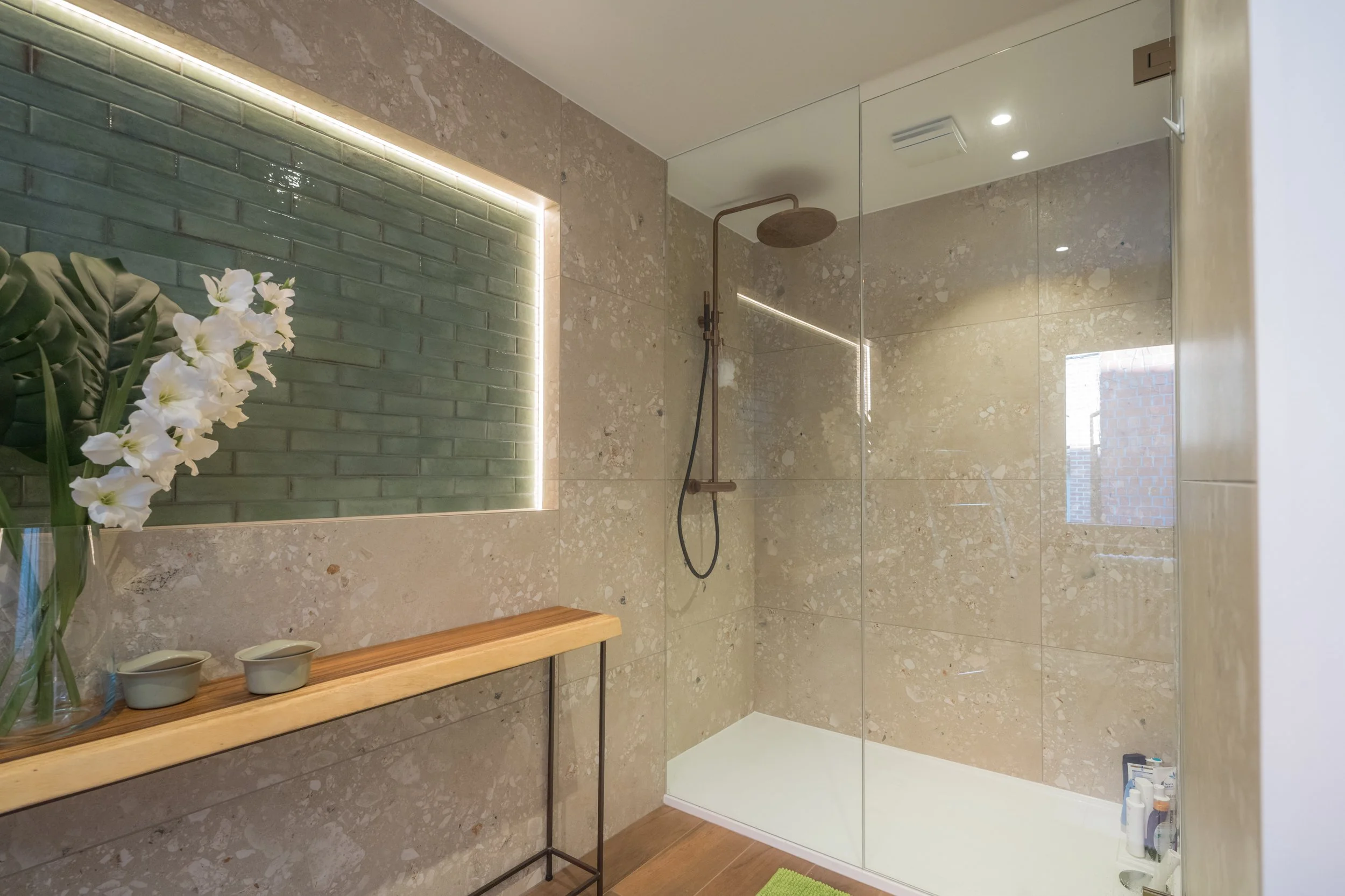 Moderne badkamer met inloopdouche, groene bakstenen muur, bloemen en spiegellamp.