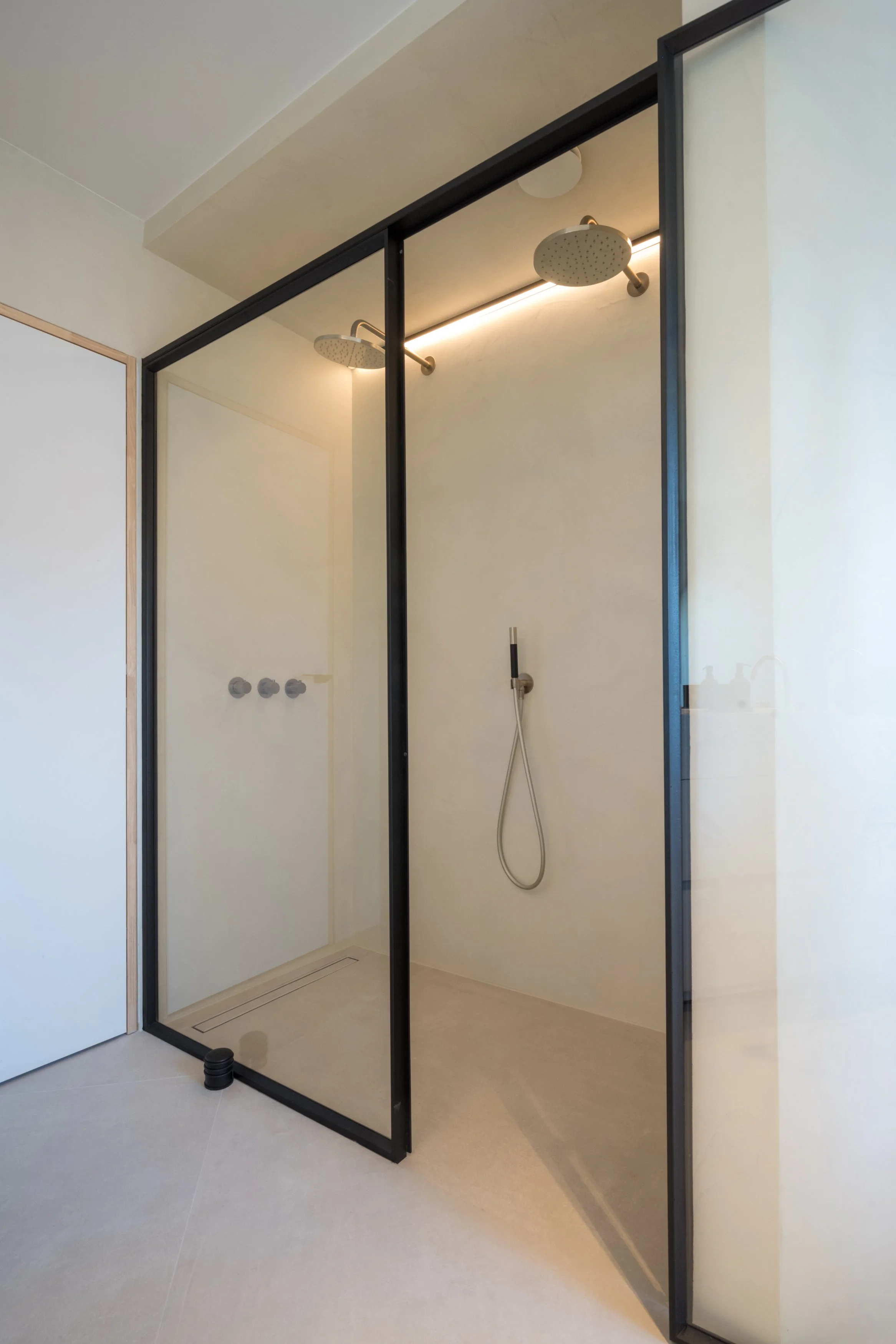 Een moderne inloopdouche met twee koppen, een handdouche en een glazen deur met zwarte frame.