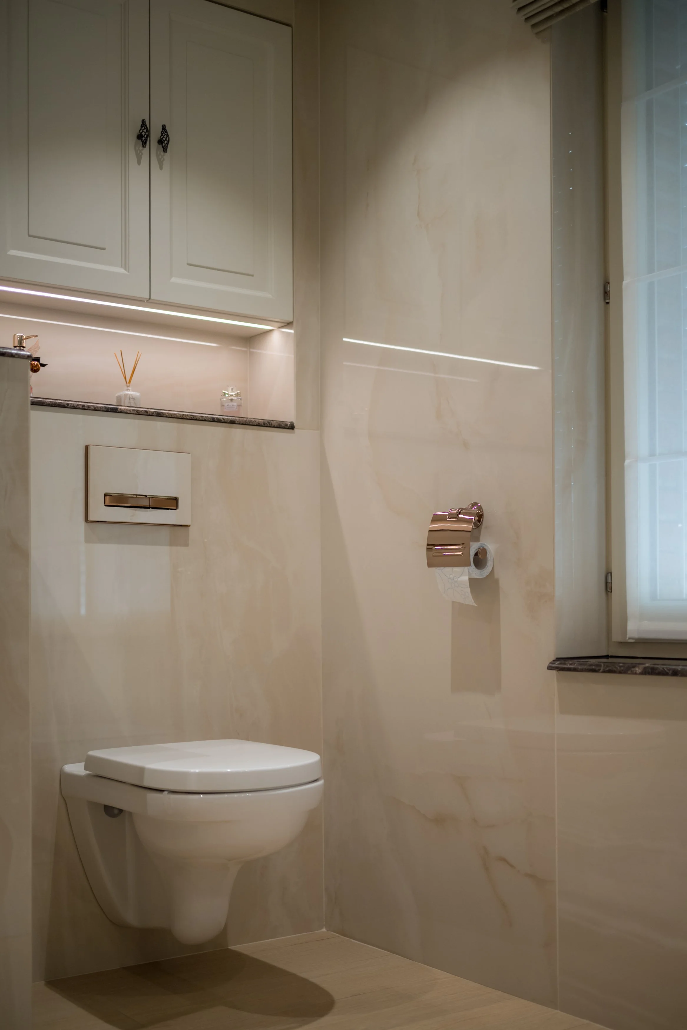 Een nette badkamer met wandtoilet, een venster met shutters, en beige marmerachtige tegels.