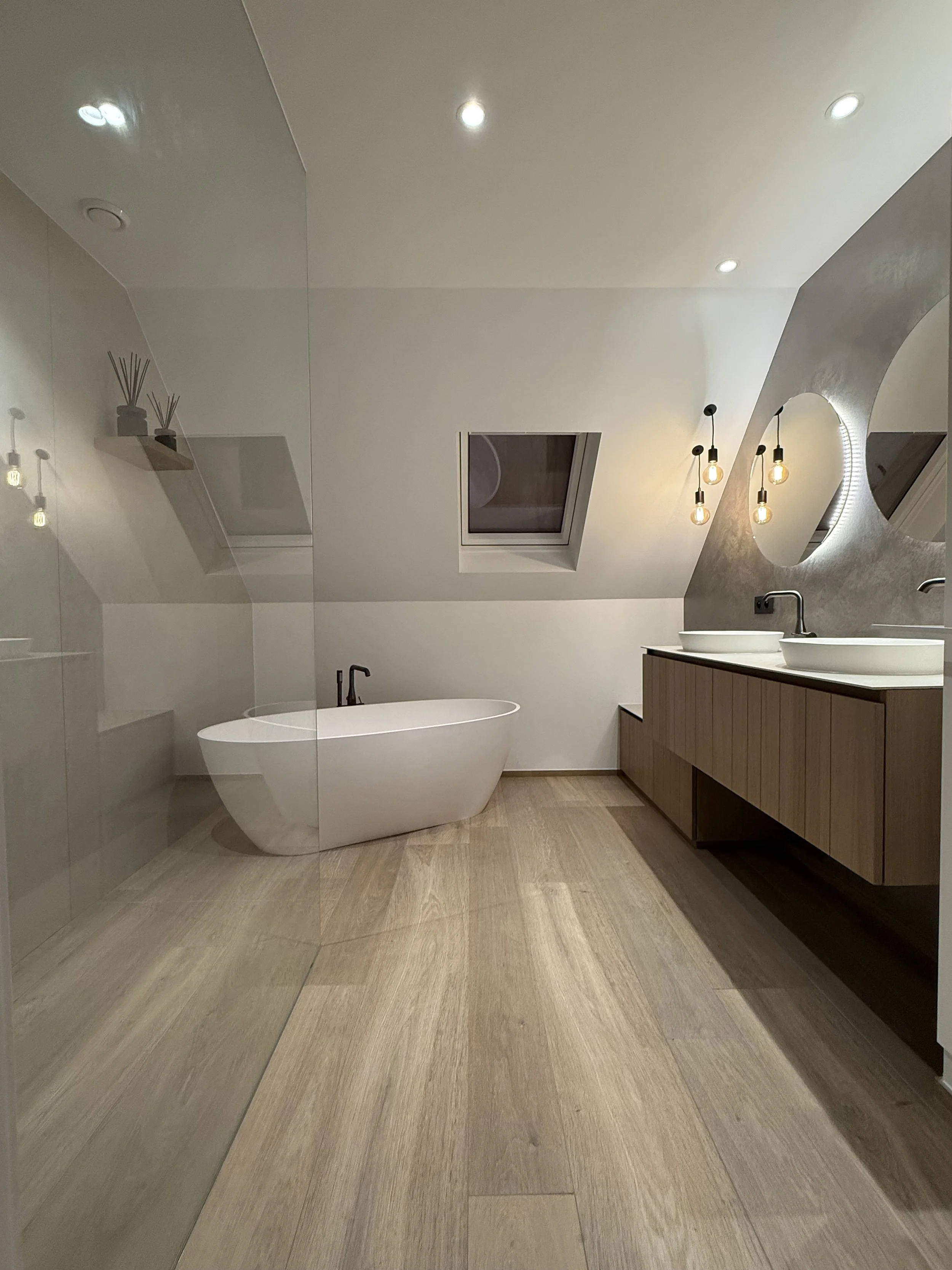 Moderne badkamer met vrijstaand bad, dubbel wastafel, ronde spiegel, houten kast en plafondramen, lichte houten vloer, en wandverlichting.