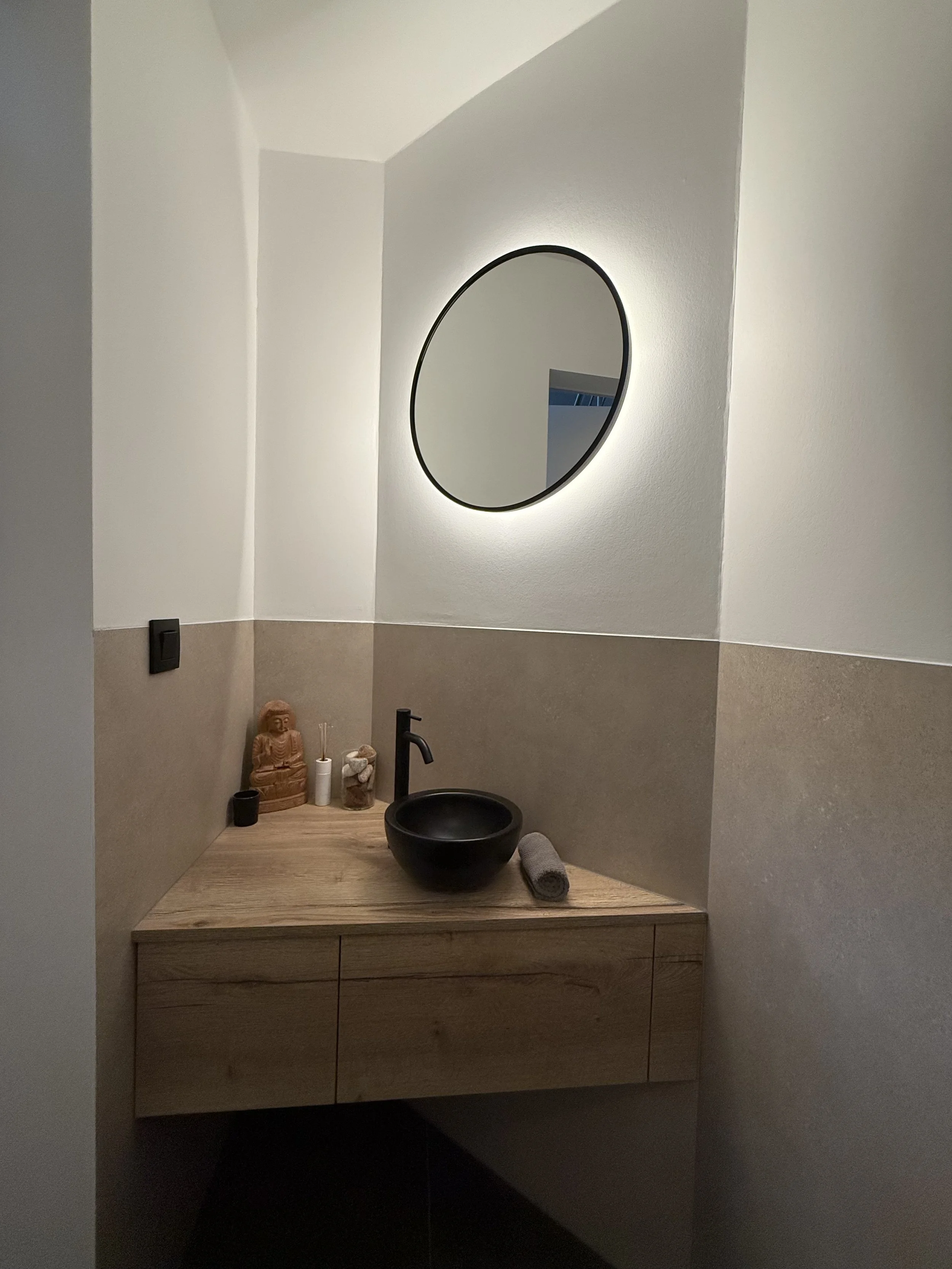 Moderne badkamer met een houten wastafel en een zwart basiswasbak, een ronde spiegel met verlichting eromheen, decoratieve objecten zoals een boeddhabeeld, een stok met geurende stokjes, een stenen schaal, en een handdoek.