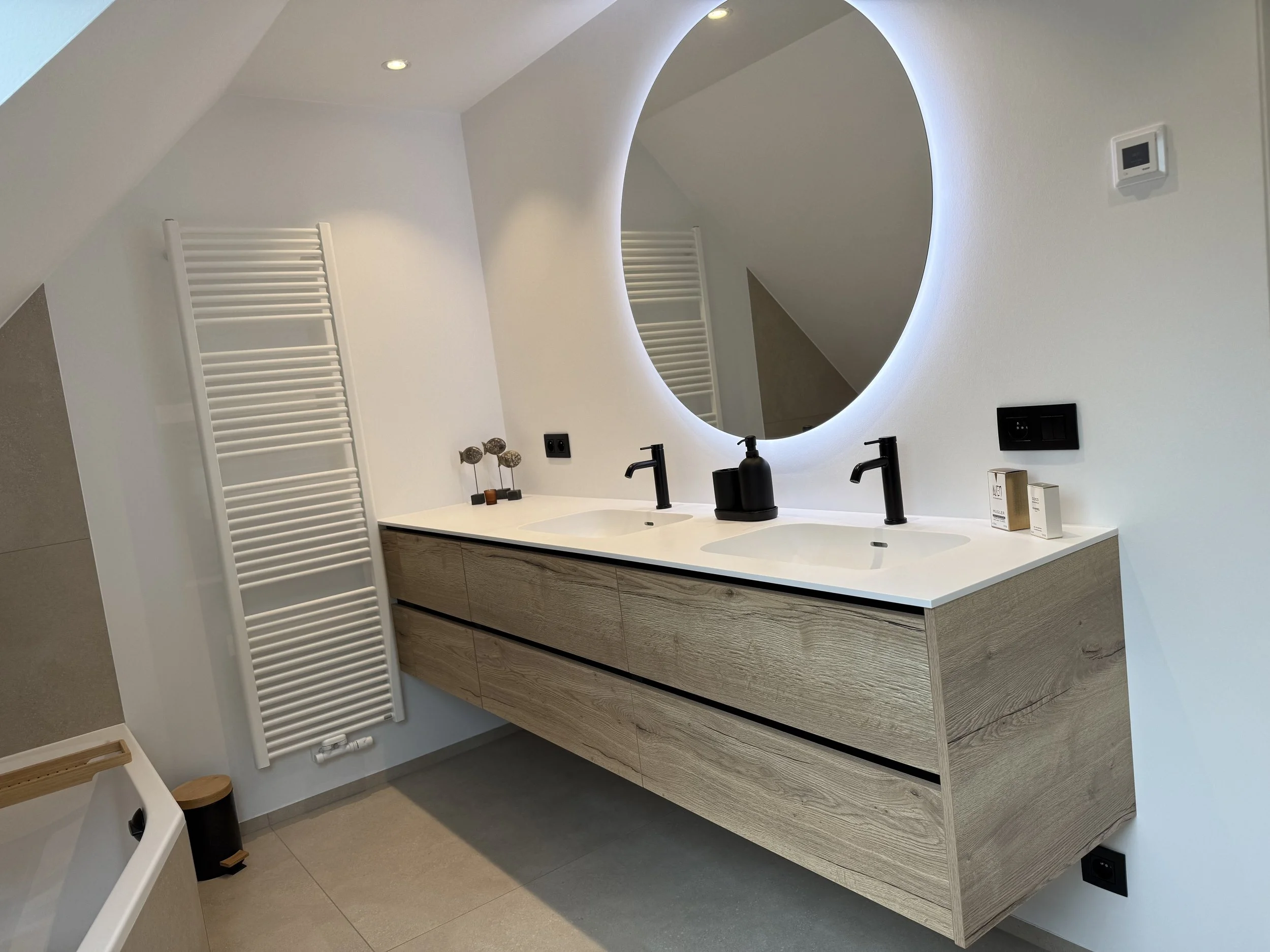 Moderne badkamer met dubbele wasbak, grote ronde verlichte spiegel, houten wastafelmeubel en ronde decoratie ornamenten.