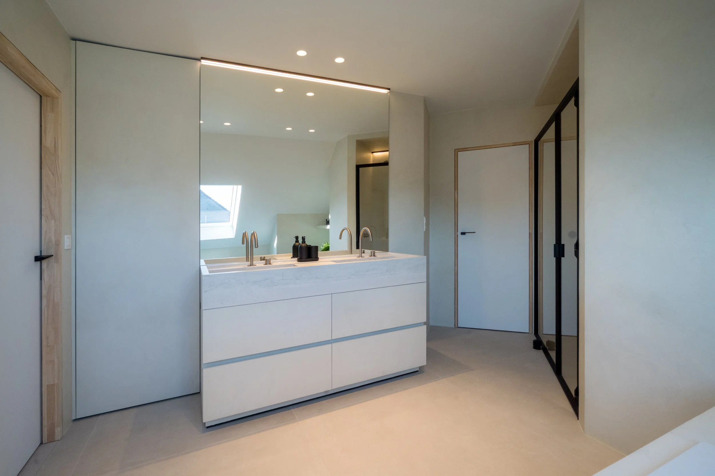 Minimalistische badkamer met wit wastafelmeubel, dubbelstaand kranen, grote spiegel, zachte verlichting en een raam voor natuurlijk licht.