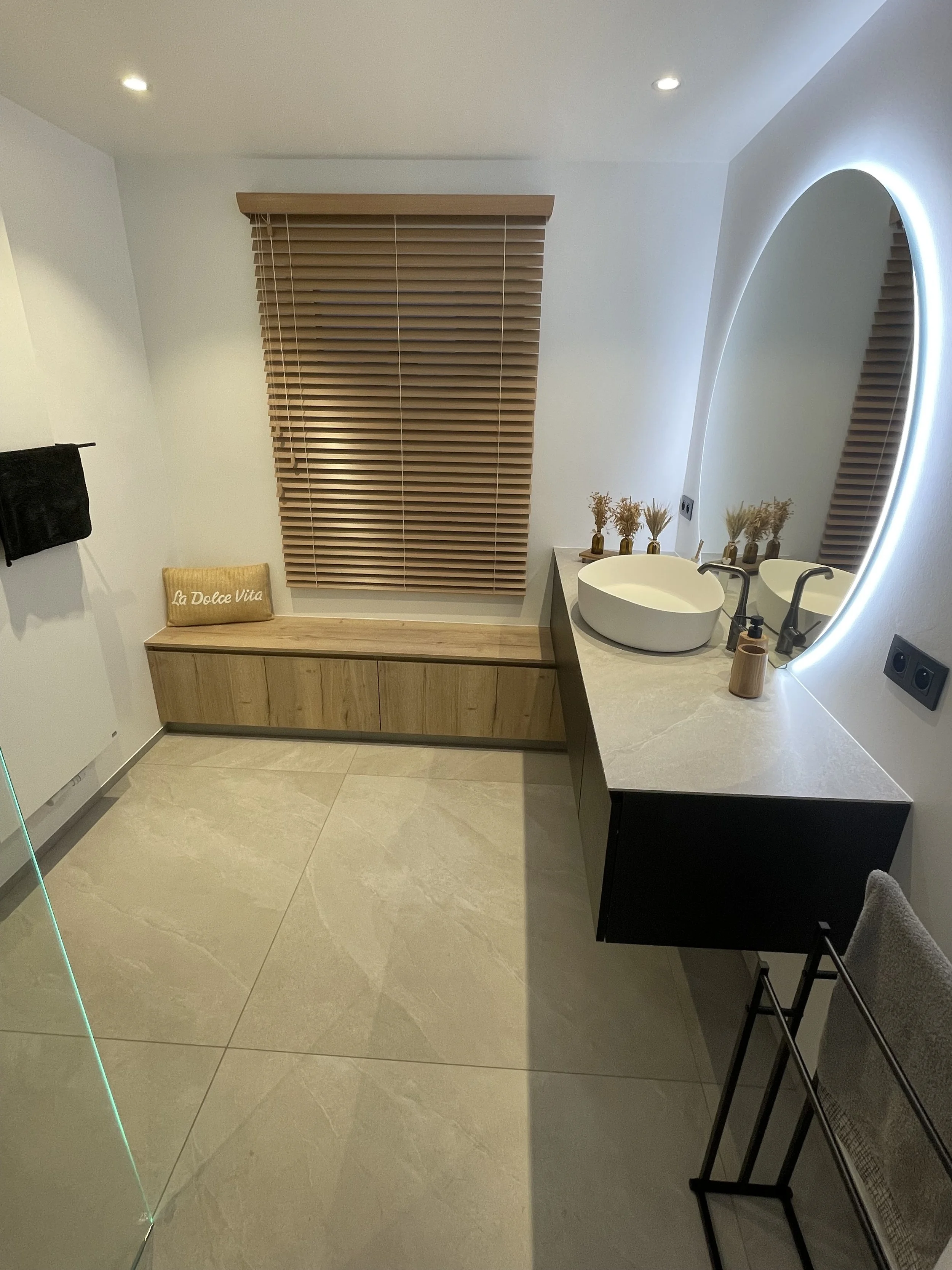 Moderne badkamer met een ronde spiegel met verlichting, een wastafel met kraan en accessoires, houten jaloezieën voor het raam, een bench onder het raam met een kussen, een handdoekhouder met handdoek en een vloer met grote lichtgekleurde tegels.