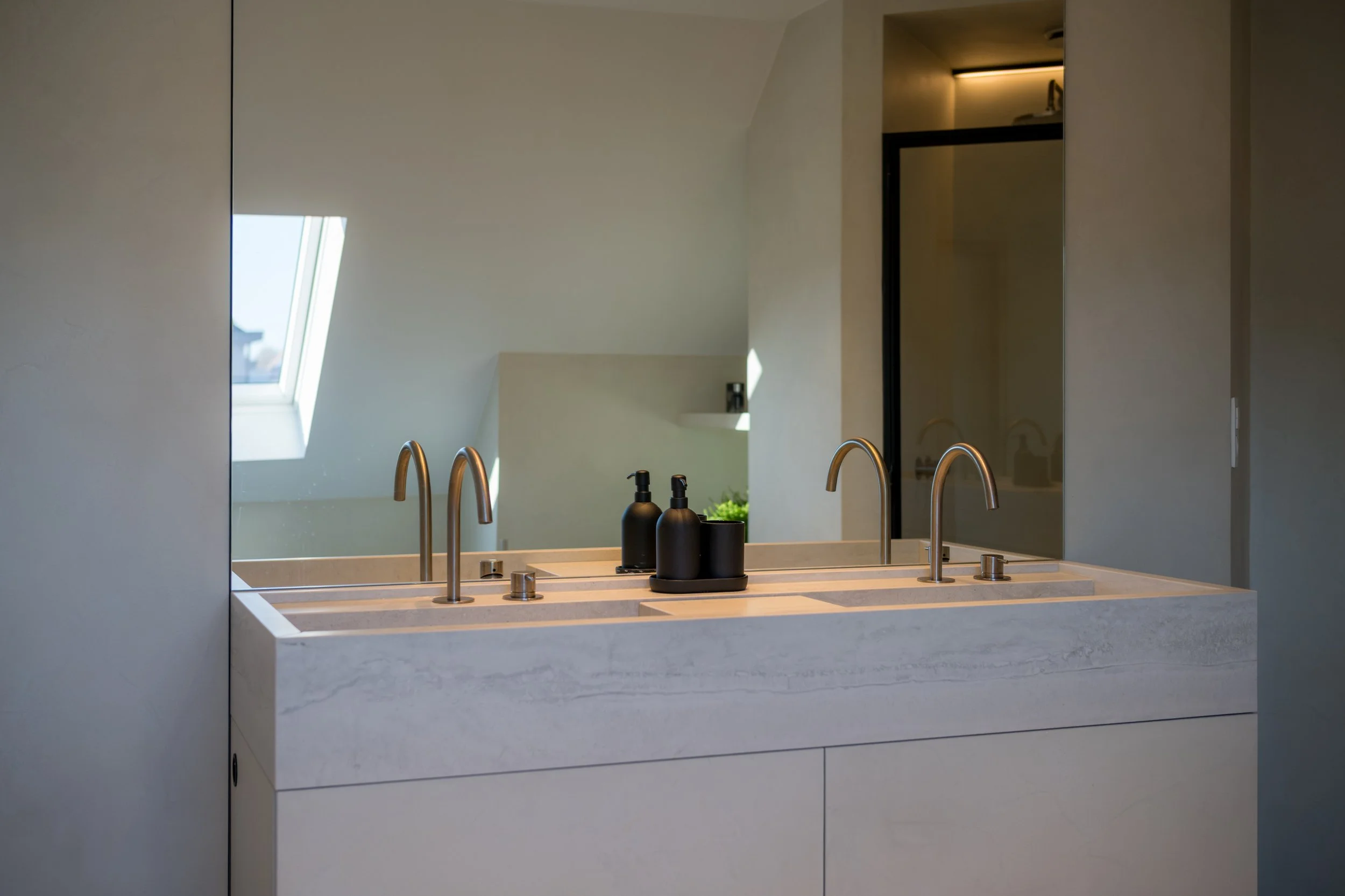 Een wit wastafelmeubel met twee kranen en zwarte zeepdispensers, tegen een grote spiegel in een modern badkamer met een dakraam.