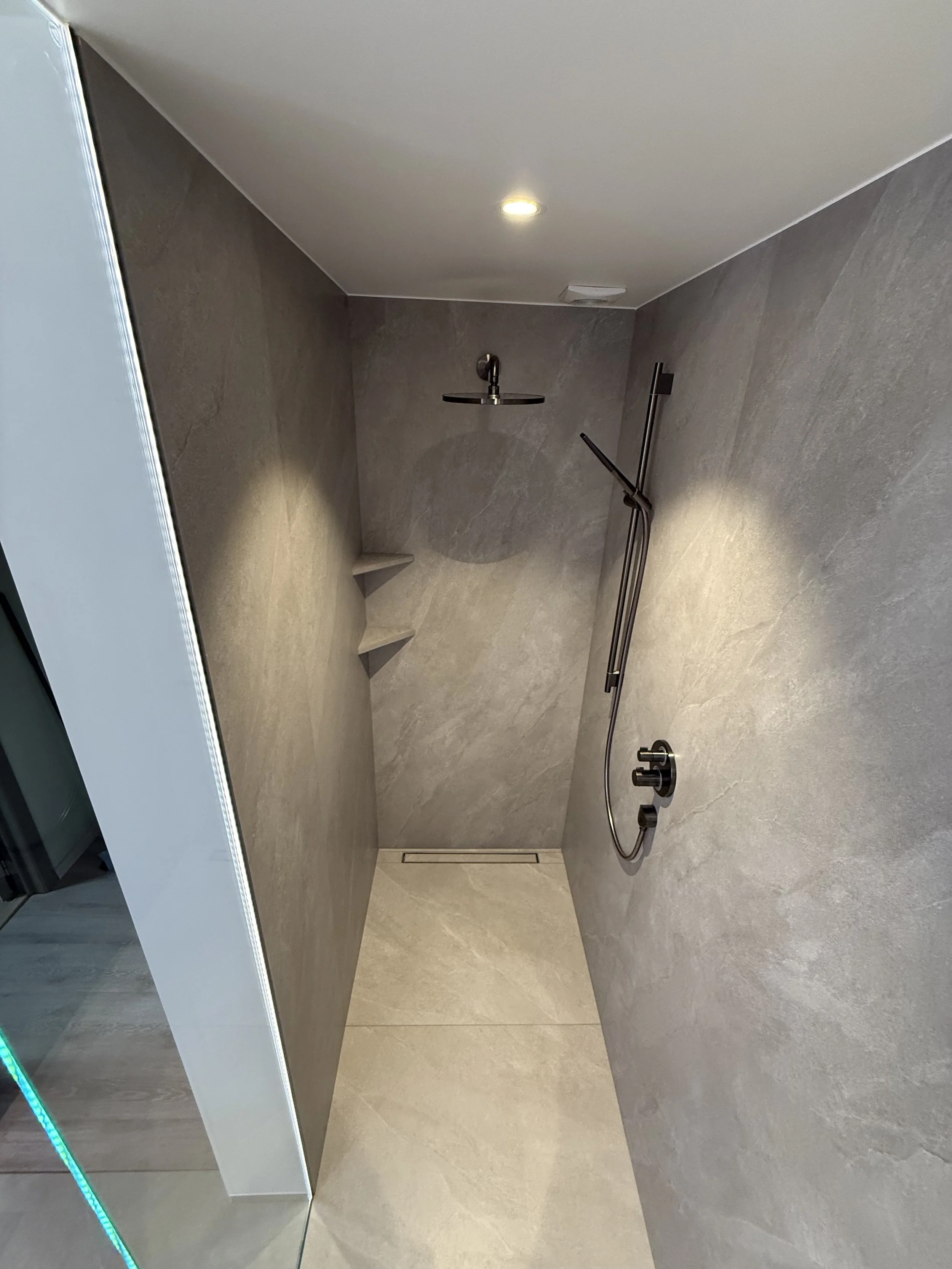 Een moderne inloopdouche met grijze tegels, een thermostaatkraan, een regenachtige douchekop en stenen ingebouwde planken.