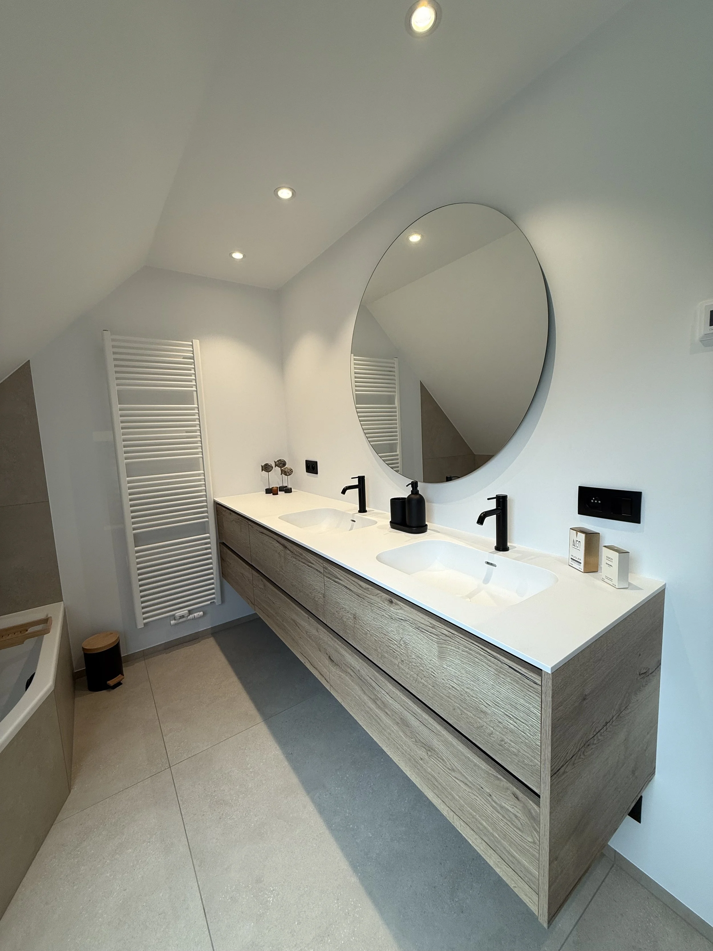 Moderne badkamer met dubbele wastafel, grote ronde spiegel, witte kachel, en zwarte accessoires.