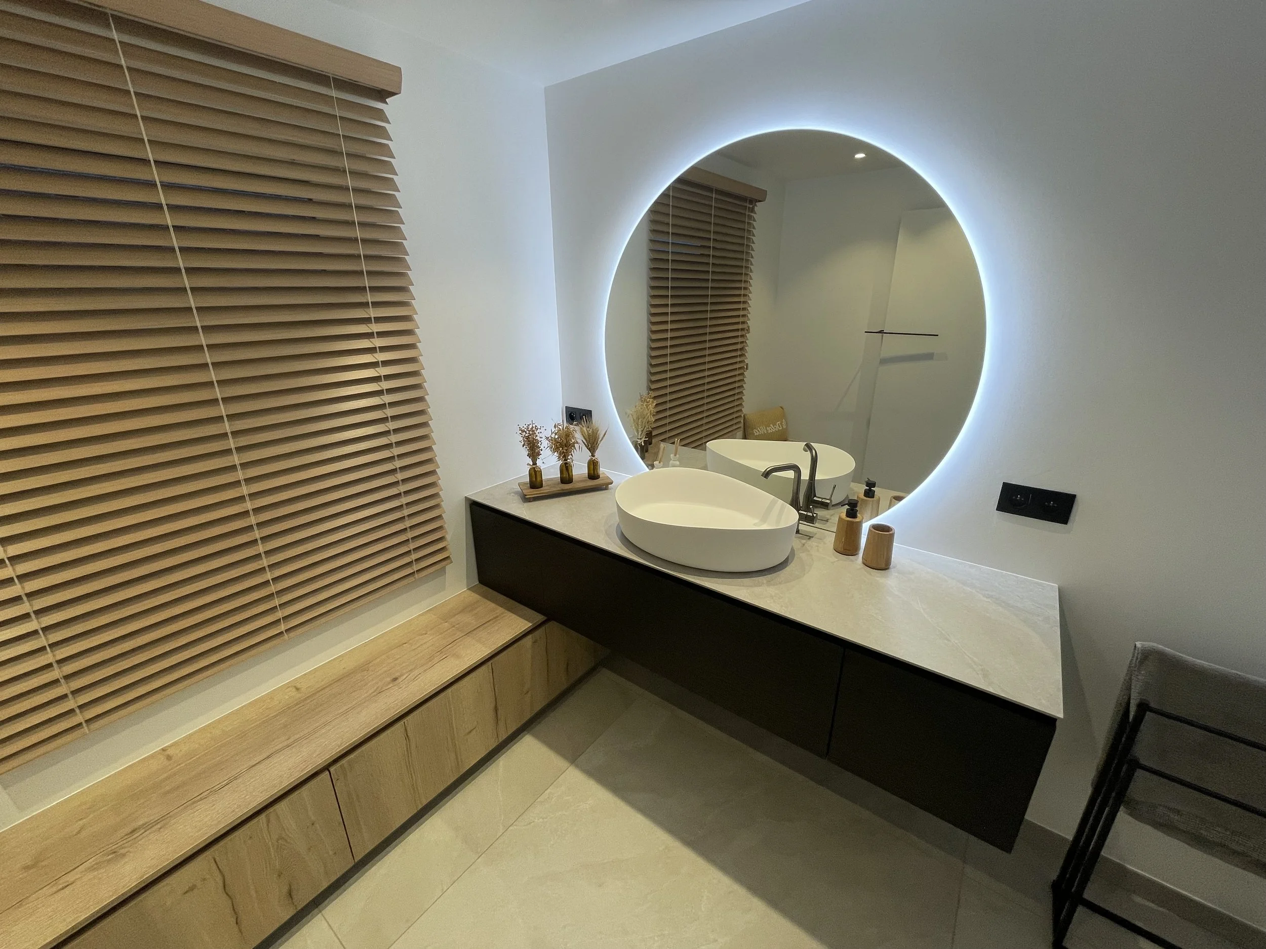 Moderne badkamer met een ronde verlichte spiegel, een witte wastafel, houten vensterblinds en een zwarte badmeubel.
