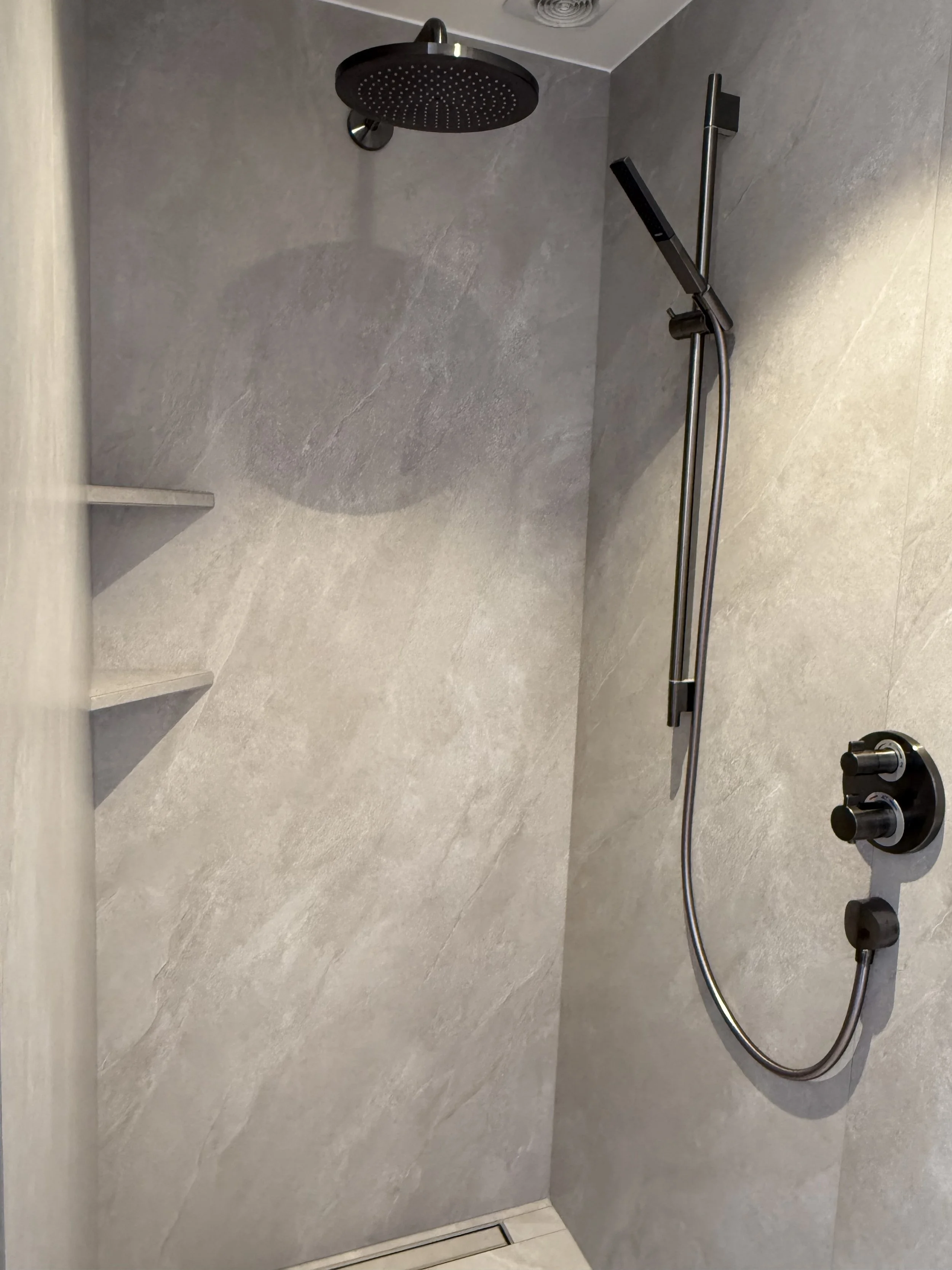 Moderne inloopdouche met een rainshower en een handdouche, betegeld met grote grijze tegels, inclusief een kleine muur met drie uitschuifbare planken.