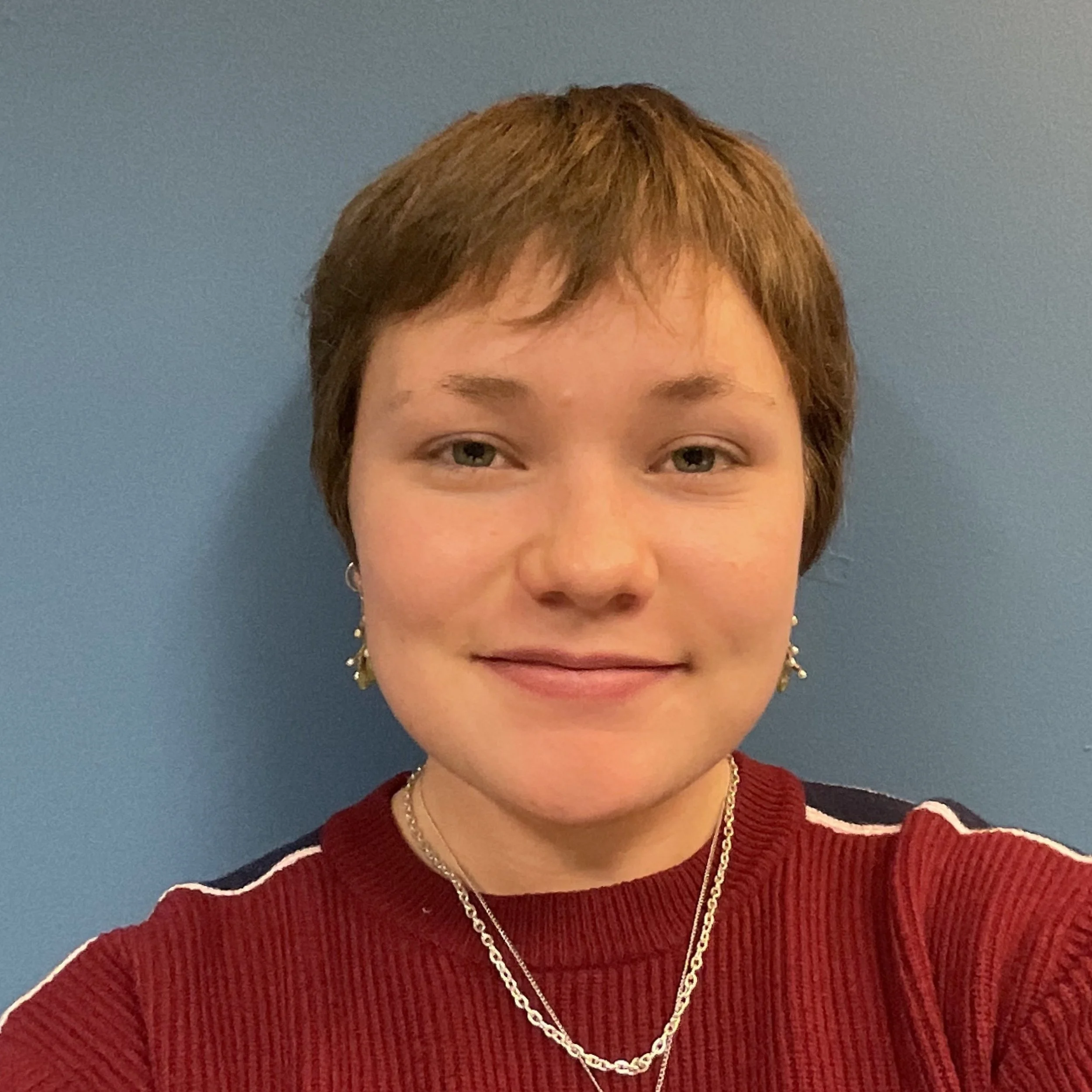 Sara has short brown hair and is wearing a red top and jewelry