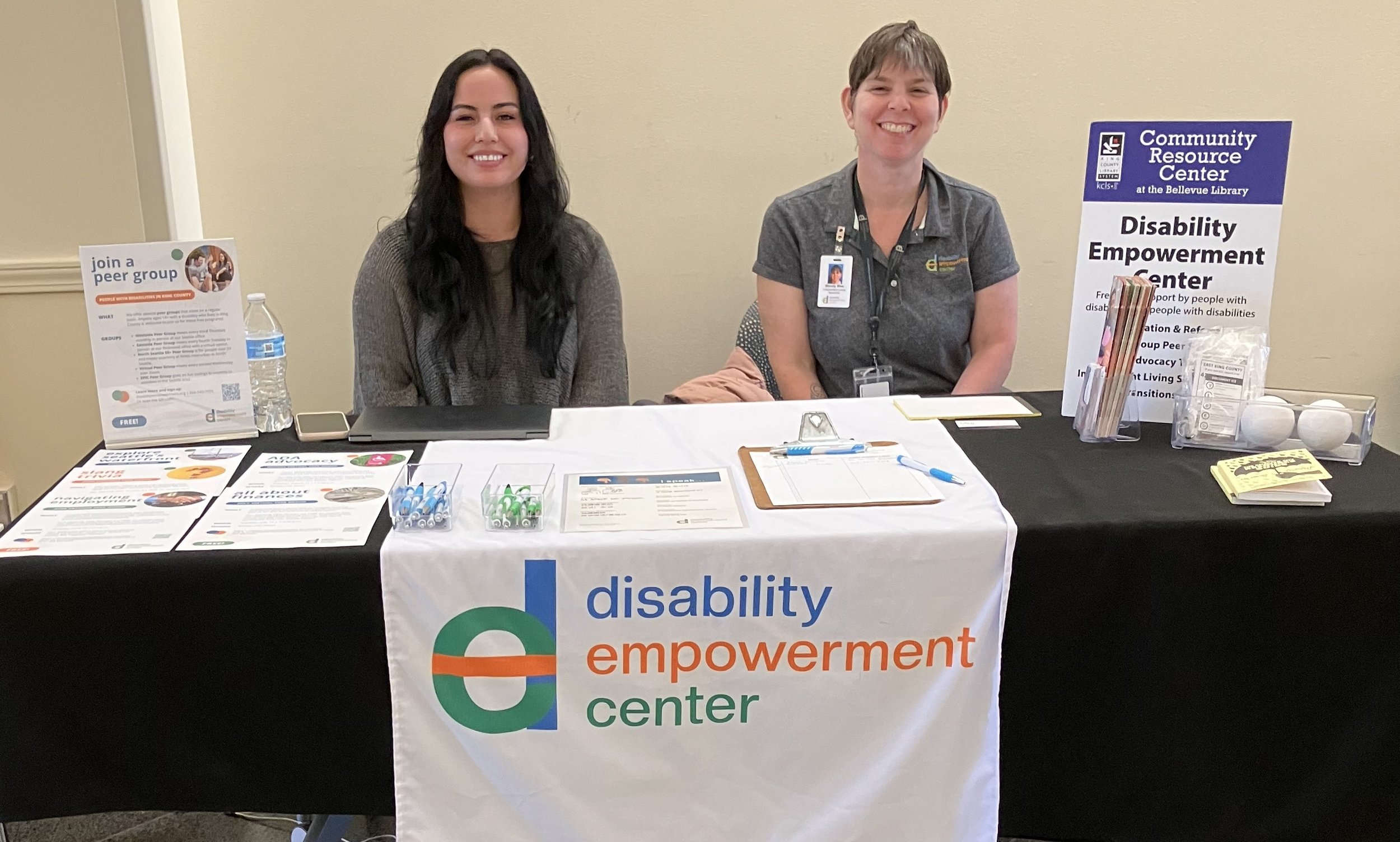 Photo of Klarysa and Mandy at a tabling event sitting smiling at the camera with resources and free giveaways in front of them