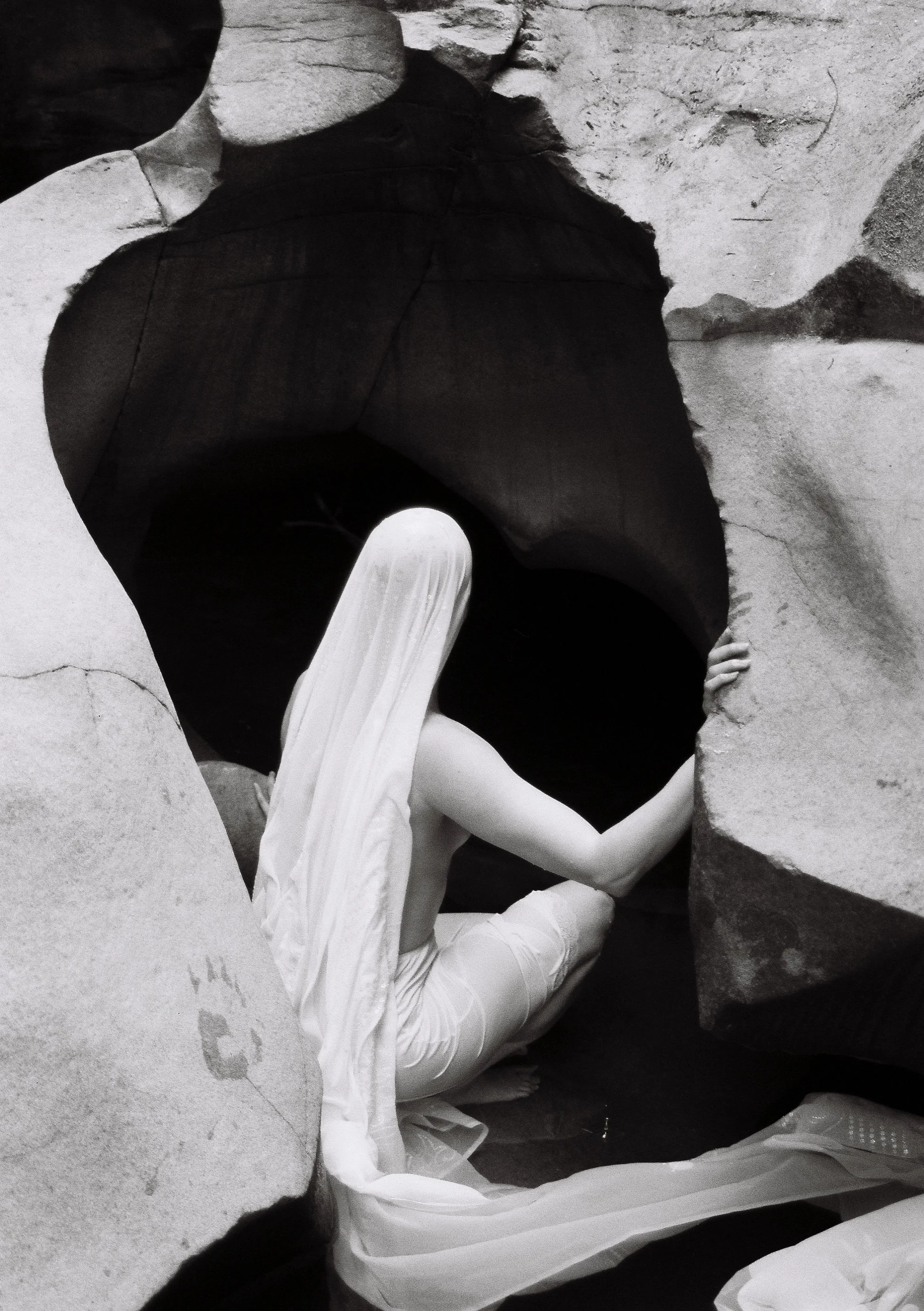 A woman in black and white holding a rock, looking into the darkness of rocks wearing a long white head scarf.