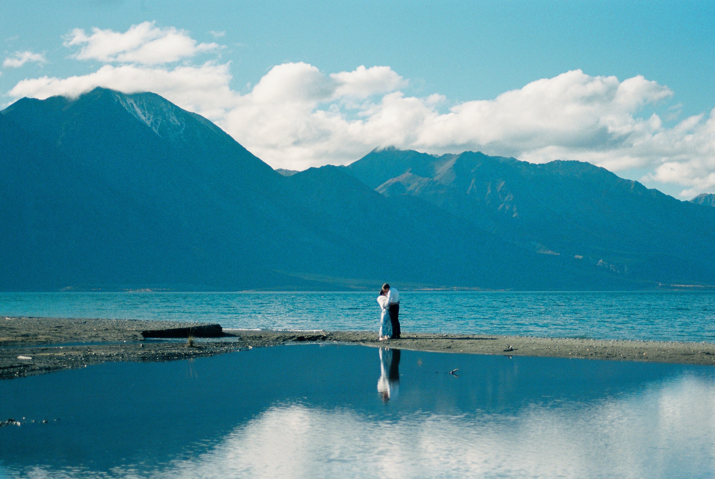 Bride and groom embracing in front of a lake with a blue mountain reflection.