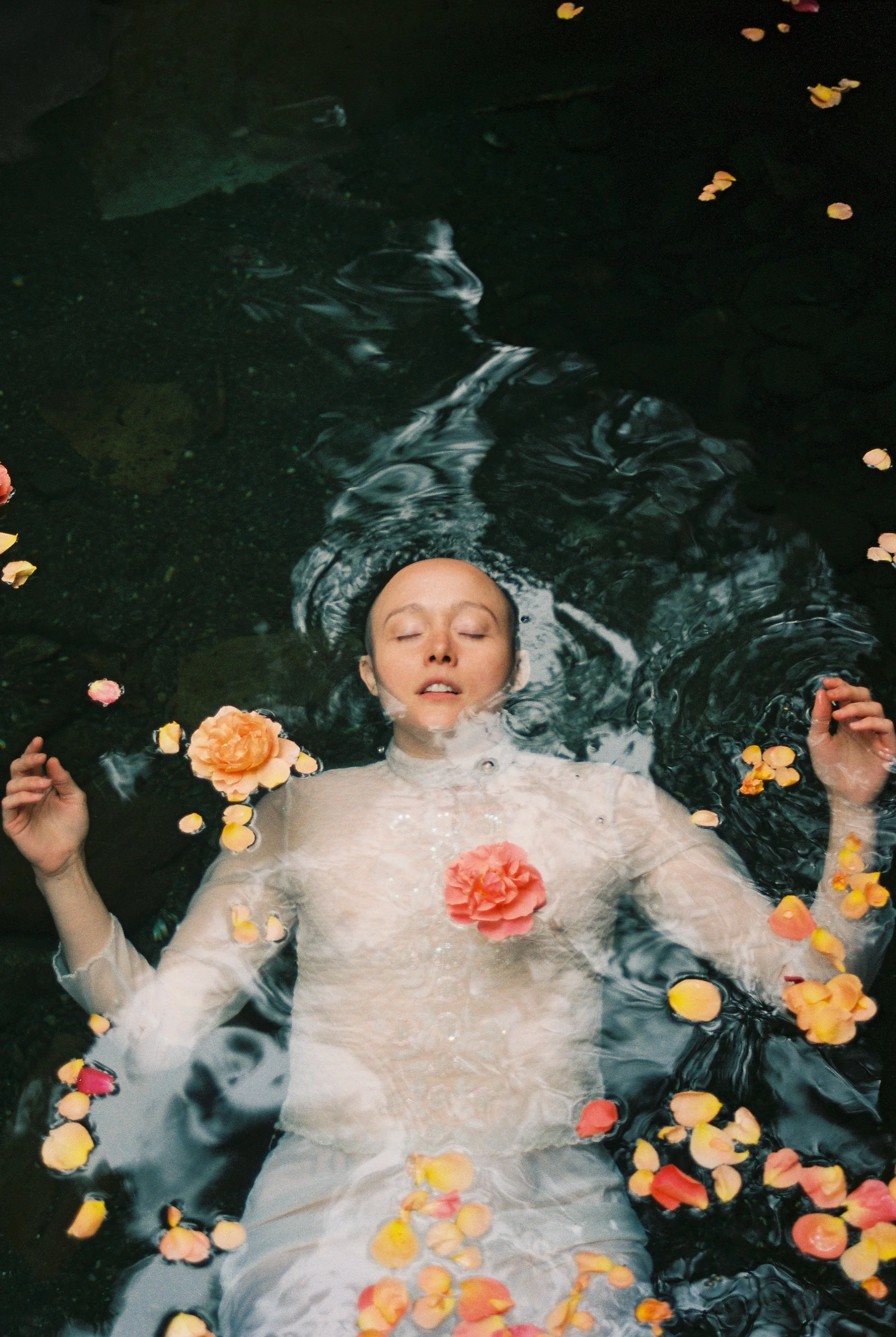 A woman lying calmly in water with flowers and petals floating around her.