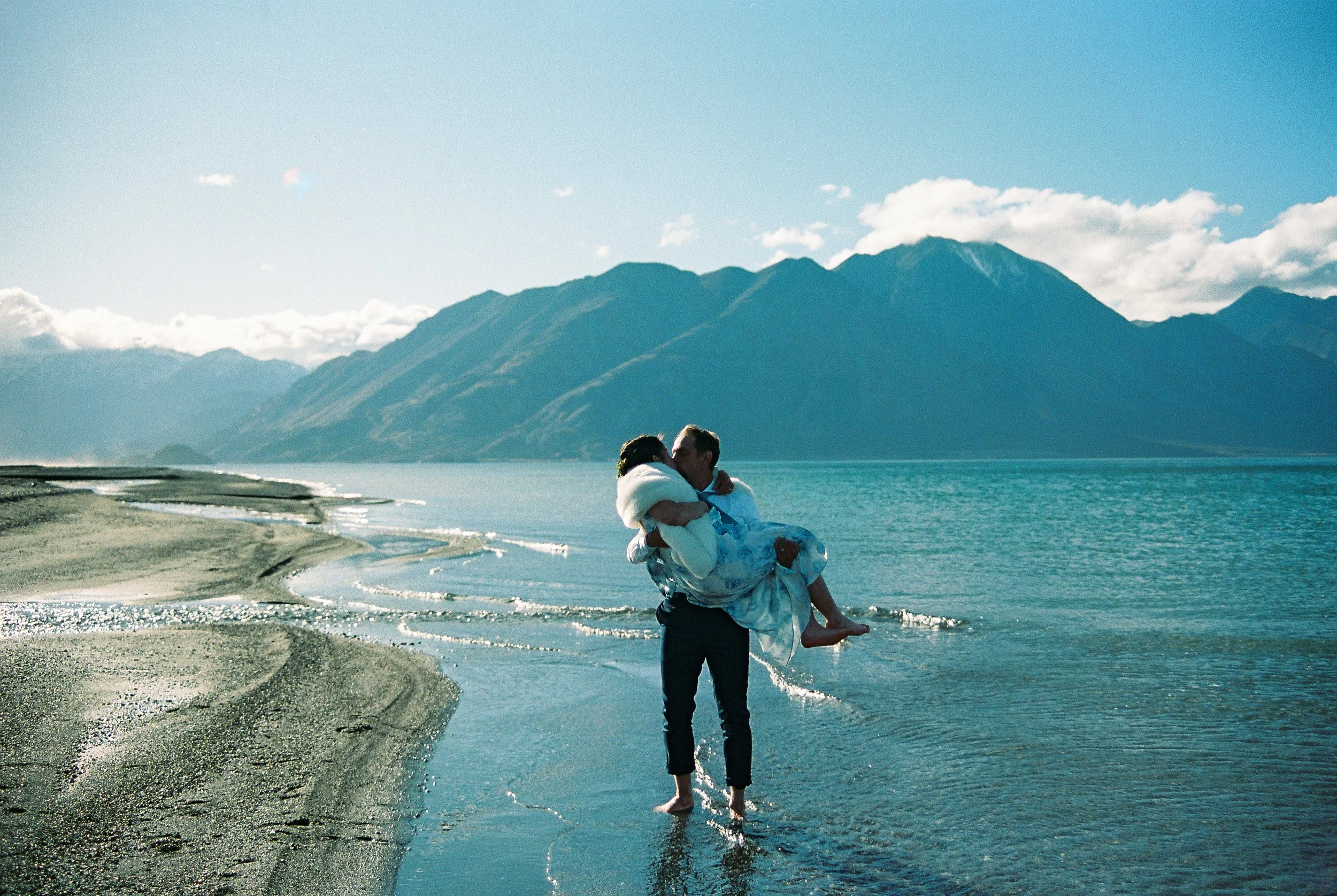 Groom holding his wife, kissing, while in a lake with blue mountains behind them.