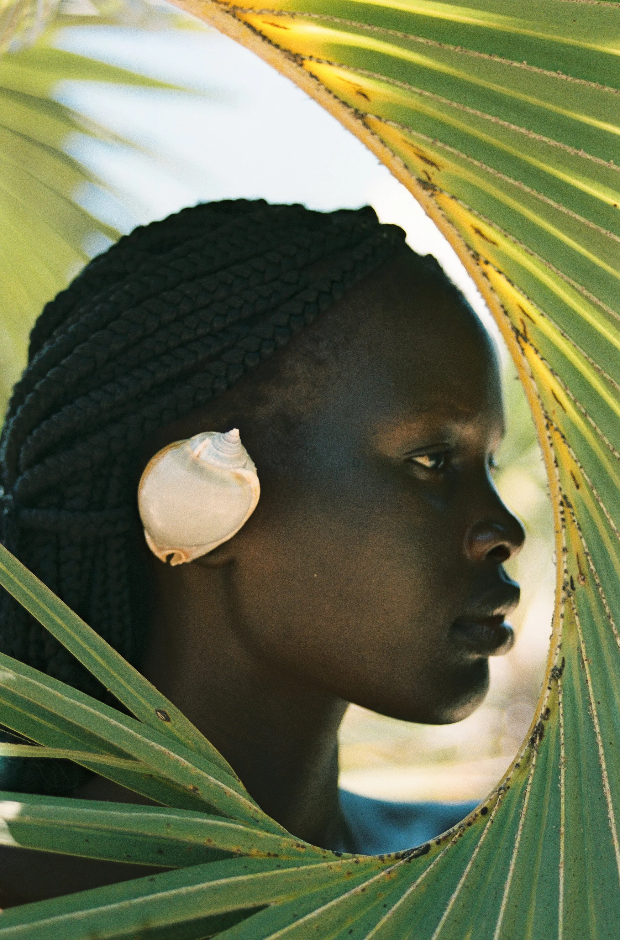 A woman with a seashell on her ear looking towards a palm leaf.